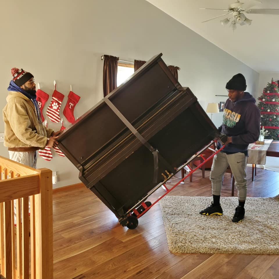 Two people wearing beanies and winter clothing use a moving dolly to transport an upright piano across a wooden floor.