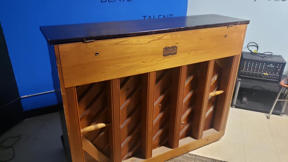 The back of a wooden upright piano, showing the structural beams, standing in a room with a blue wall and audio equipment.