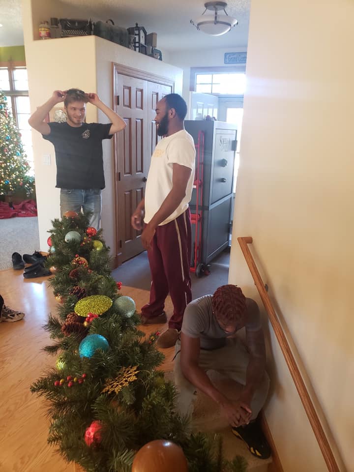 Three people inside a house near a decorated Christmas garland, with one person kneeling by a stair railing.
