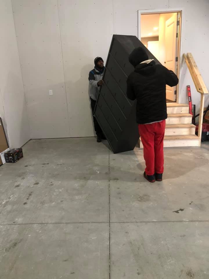 Two people in a garage moving a large, rectangular black object upright toward a set of wooden stairs.