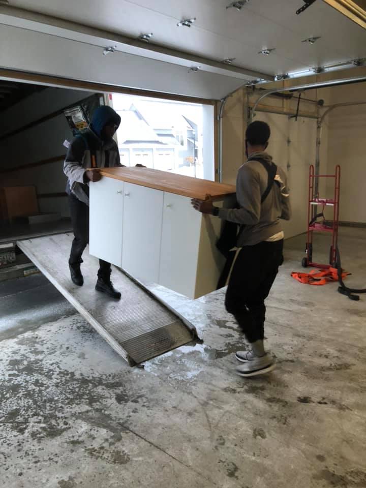 Two people carry a white cabinet with a wooden top down a truck ramp into a garage.