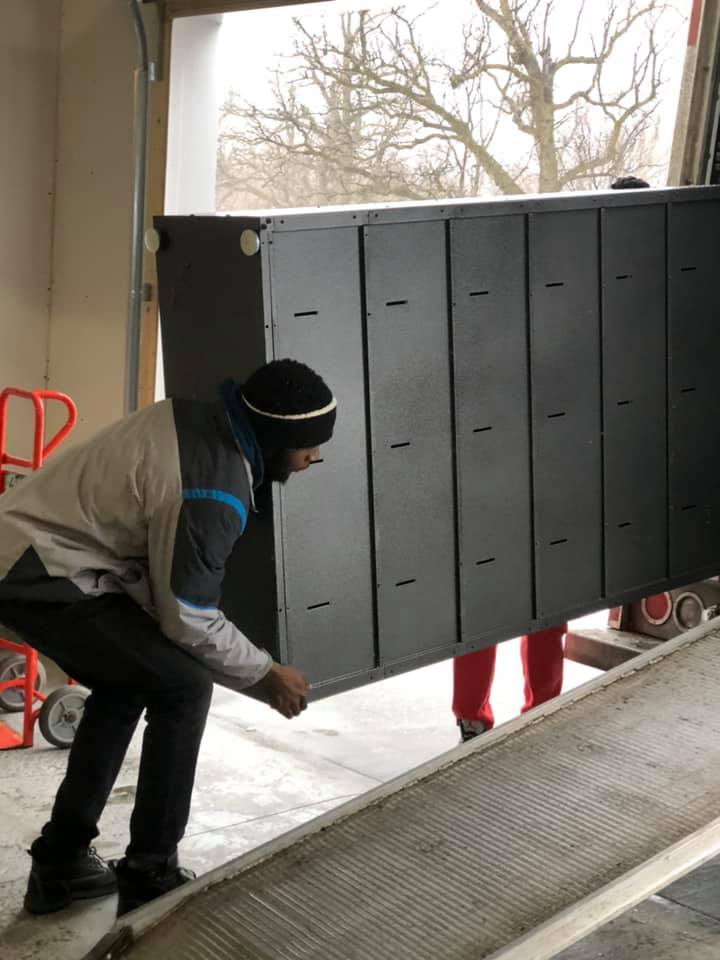 A person lifts a large black metal locker unit from a conveyor belt in a loading dock area.
