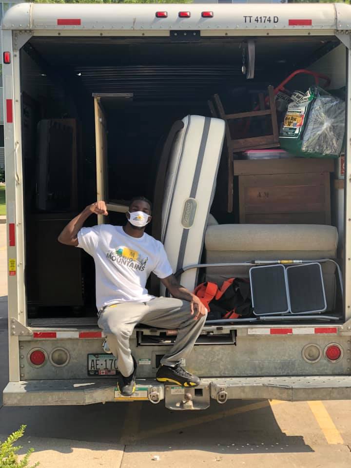 A person wearing a face mask flexes while sitting in the back of a moving truck filled with furniture and mattresses.
