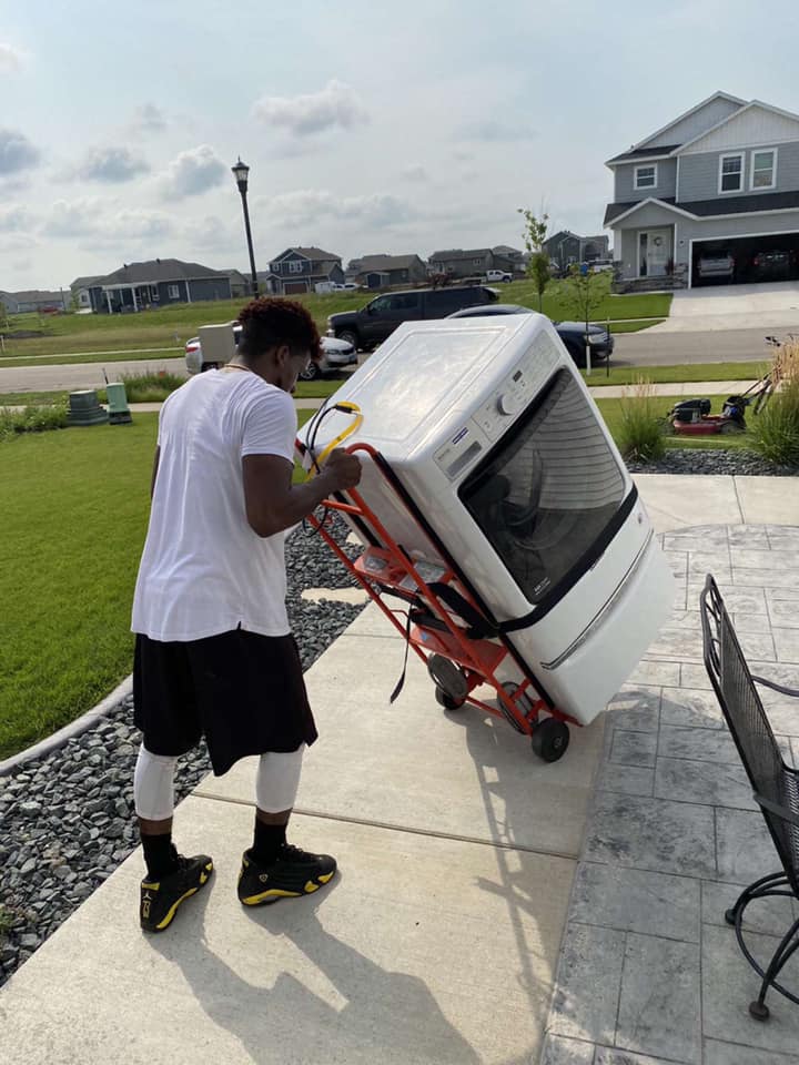 A person in a white t-shirt and dark shorts uses a hand truck to move a white dryer along a paved walkway near a house.