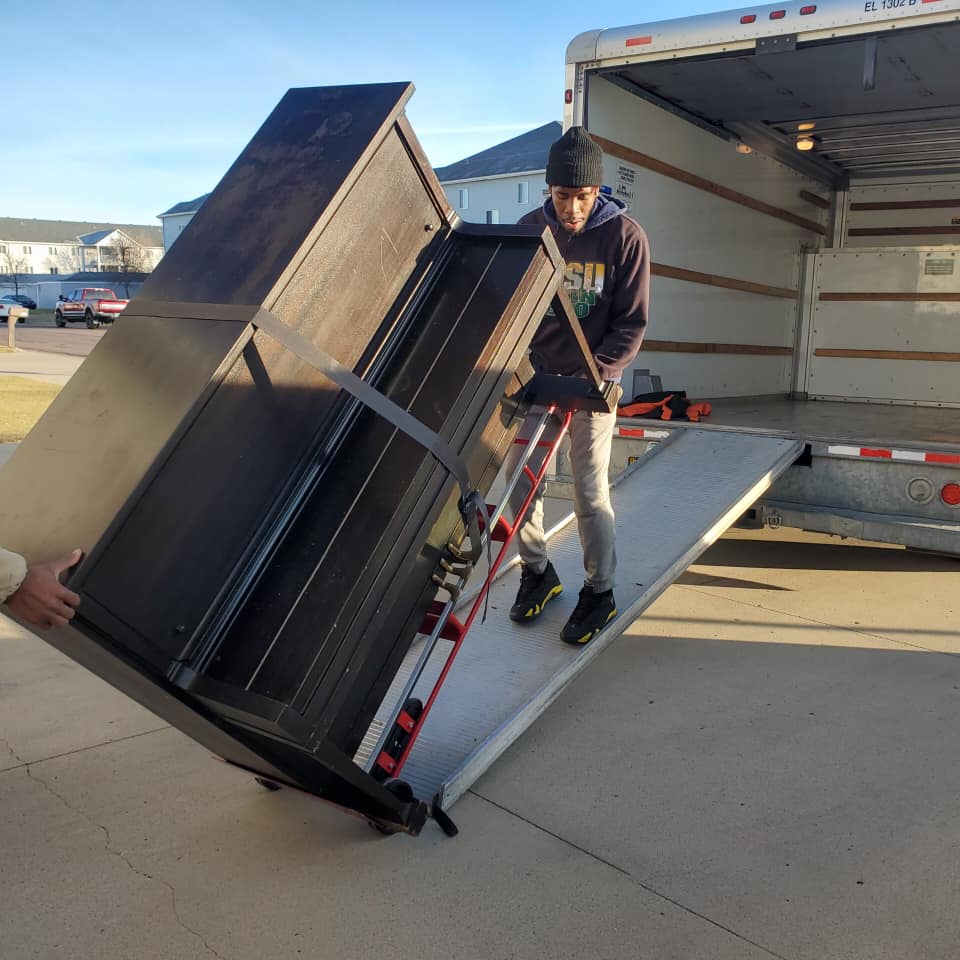 A person in a dark sweater uses a dolly to move a large, dark wooden upright piano up a truck ramp.