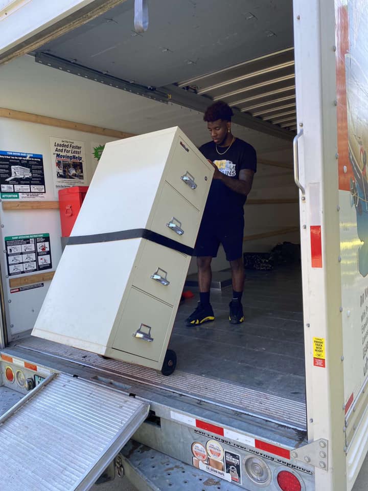 A person in a black t-shirt and shorts maneuvers a tall, beige four-drawer filing cabinet on a dolly inside a moving truck.
