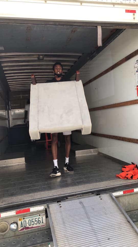 A person stands inside a moving truck, carrying a large white armchair towards the open ramp.