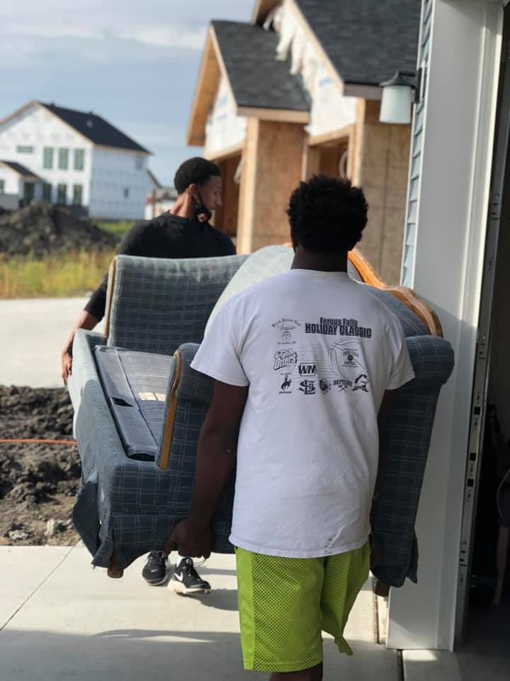 Two people carry a large gray upholstered sofa toward a residential garage under construction.