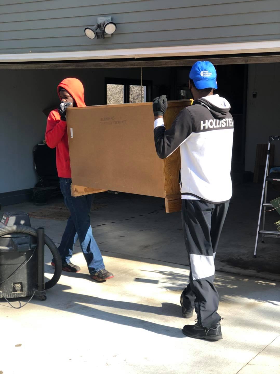 Two people carry a large, brown rectangular box from a garage into a driveway.