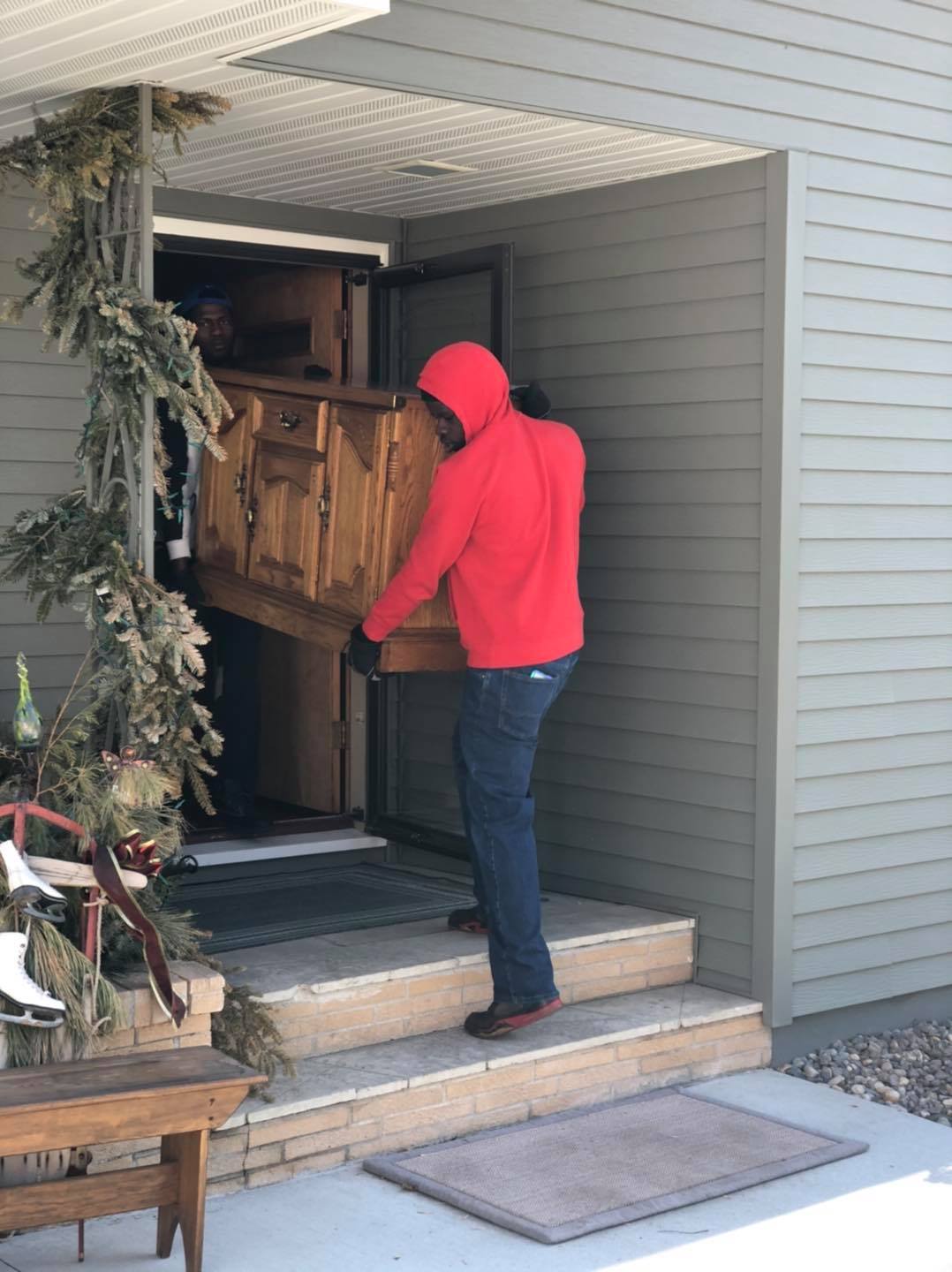 A person in a red hoodie helps carry a wooden cabinet out of the front door of a house.