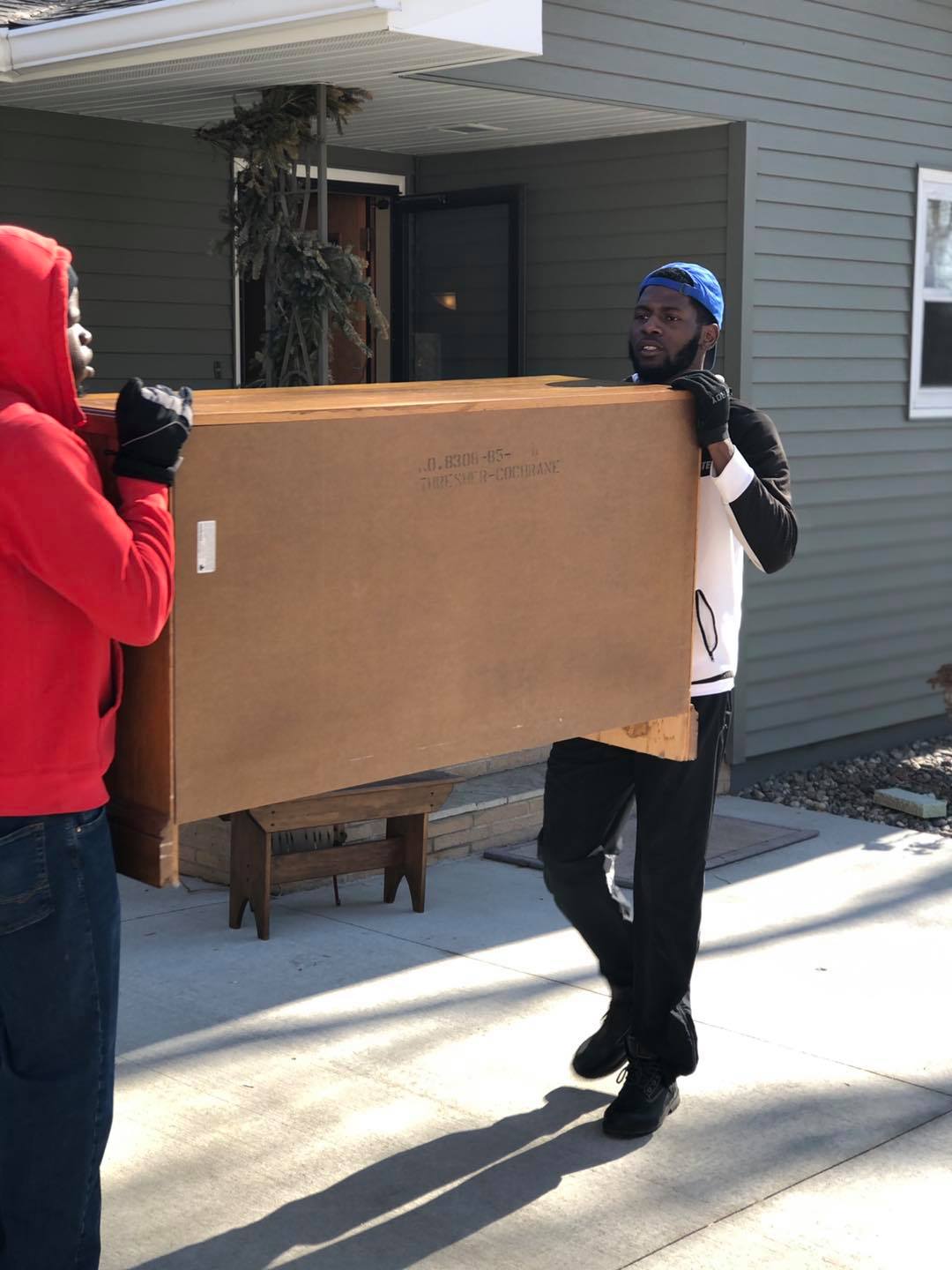 Two people wearing gloves carry a large, brown cardboard box toward the front door of a house on a sunny day.