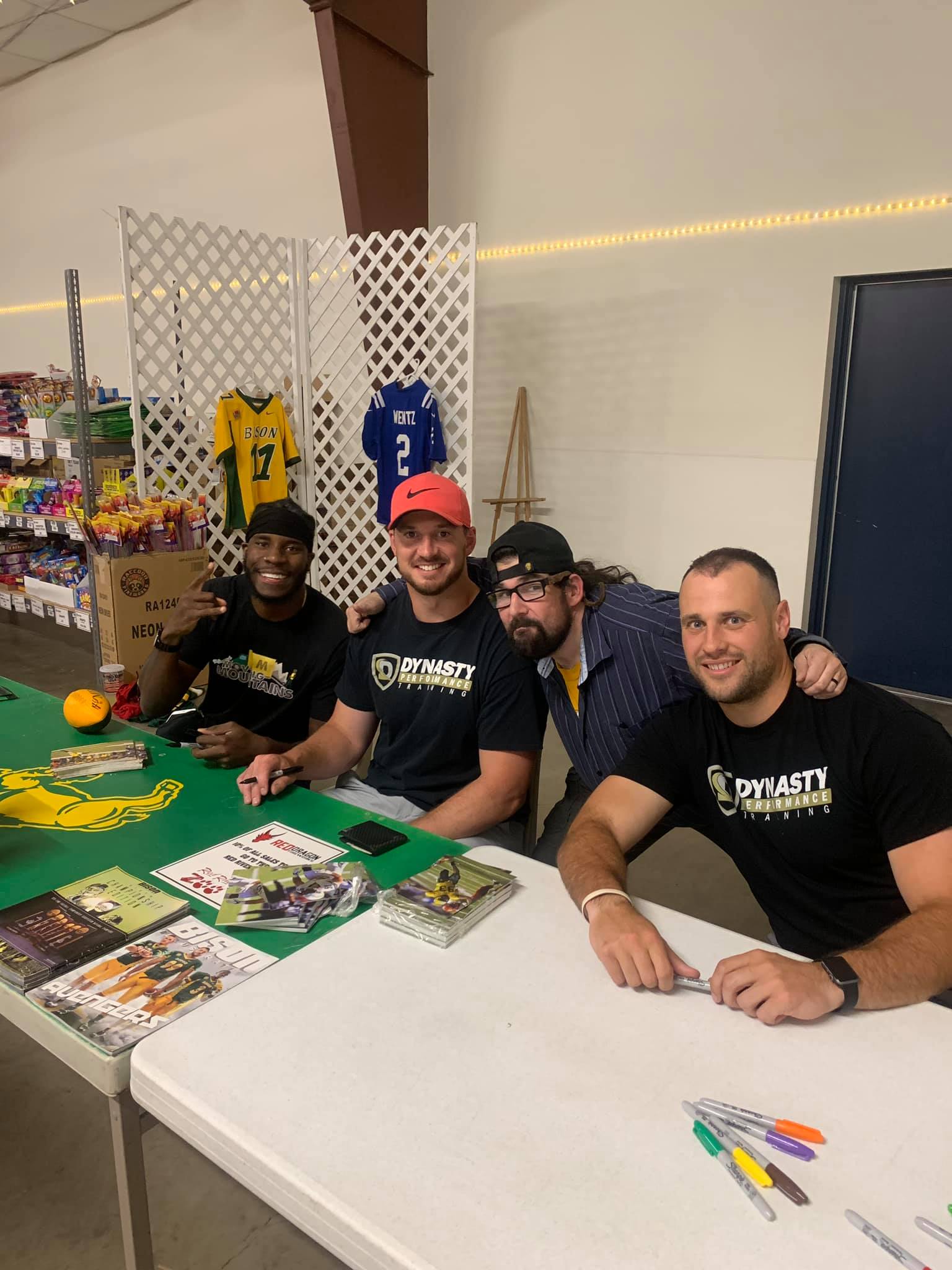 Four people sit behind a table at an autograph signing event, with sports jerseys displayed on the wall behind them.