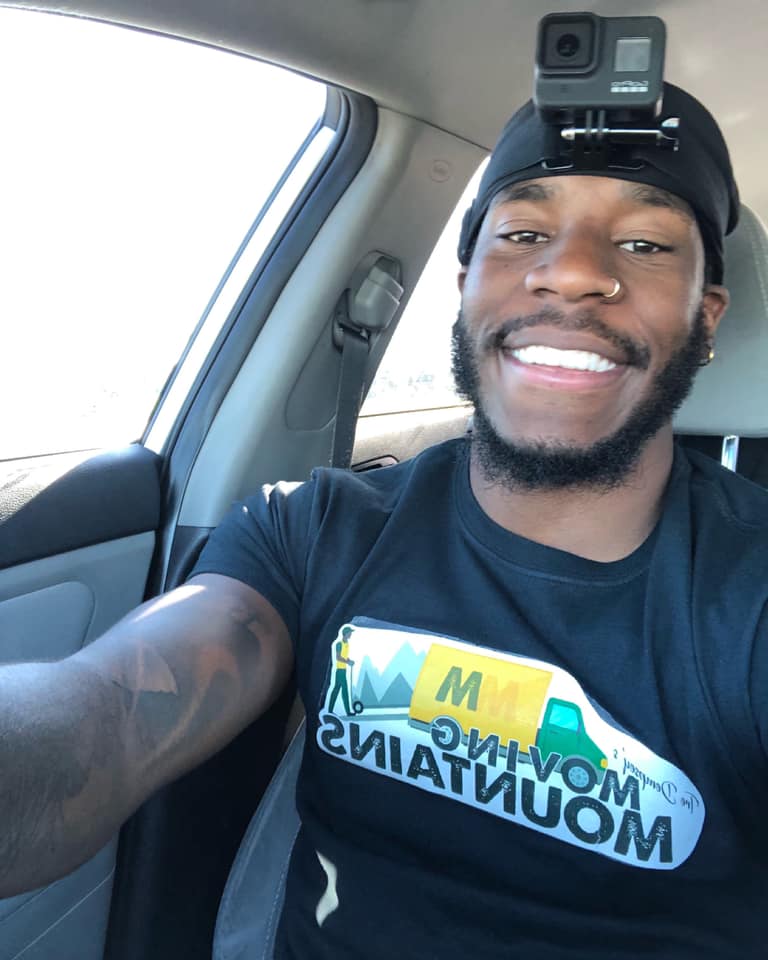 A person wearing a GoPro on their head and a Moving Mountains T-shirt smiles while seated in a car.