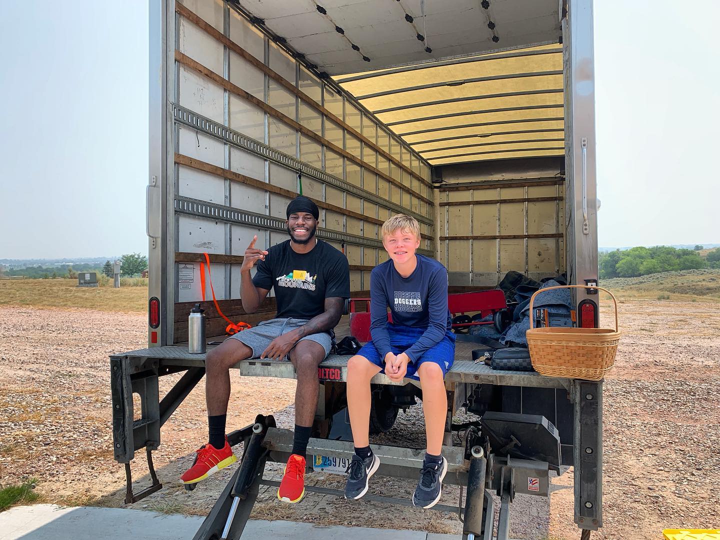 Two people sitting on the back of a large moving truck parked on a gravel lot under a hazy sky.