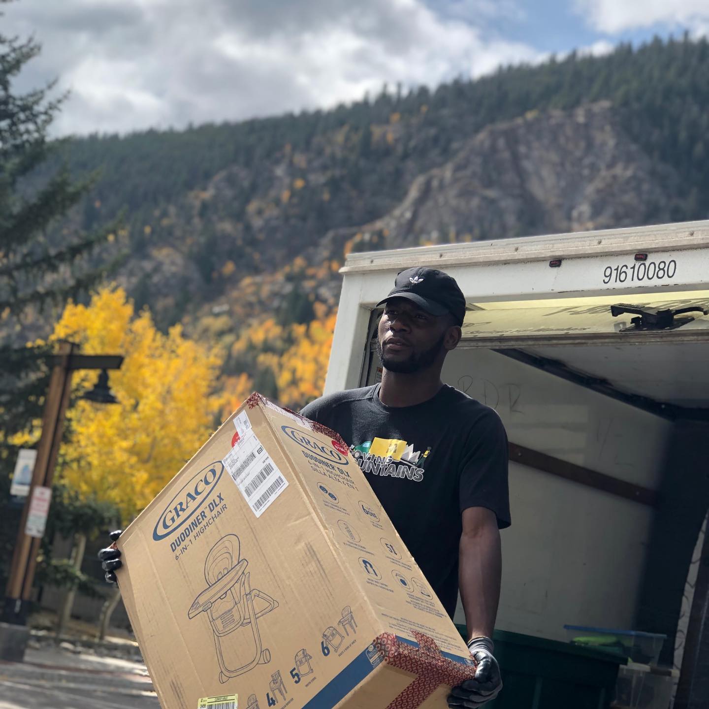 A person wearing a black cap and t-shirt carries a large Graco box out of a truck against a mountain and autumn trees.