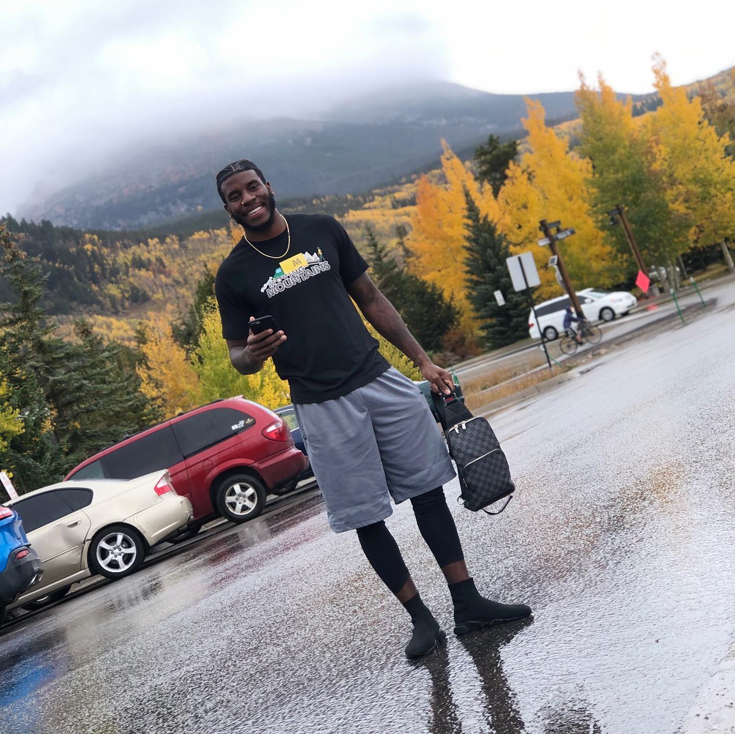A smiling person stands in a rain-slicked parking lot before autumn mountains, wearing a black t-shirt and grey shorts.