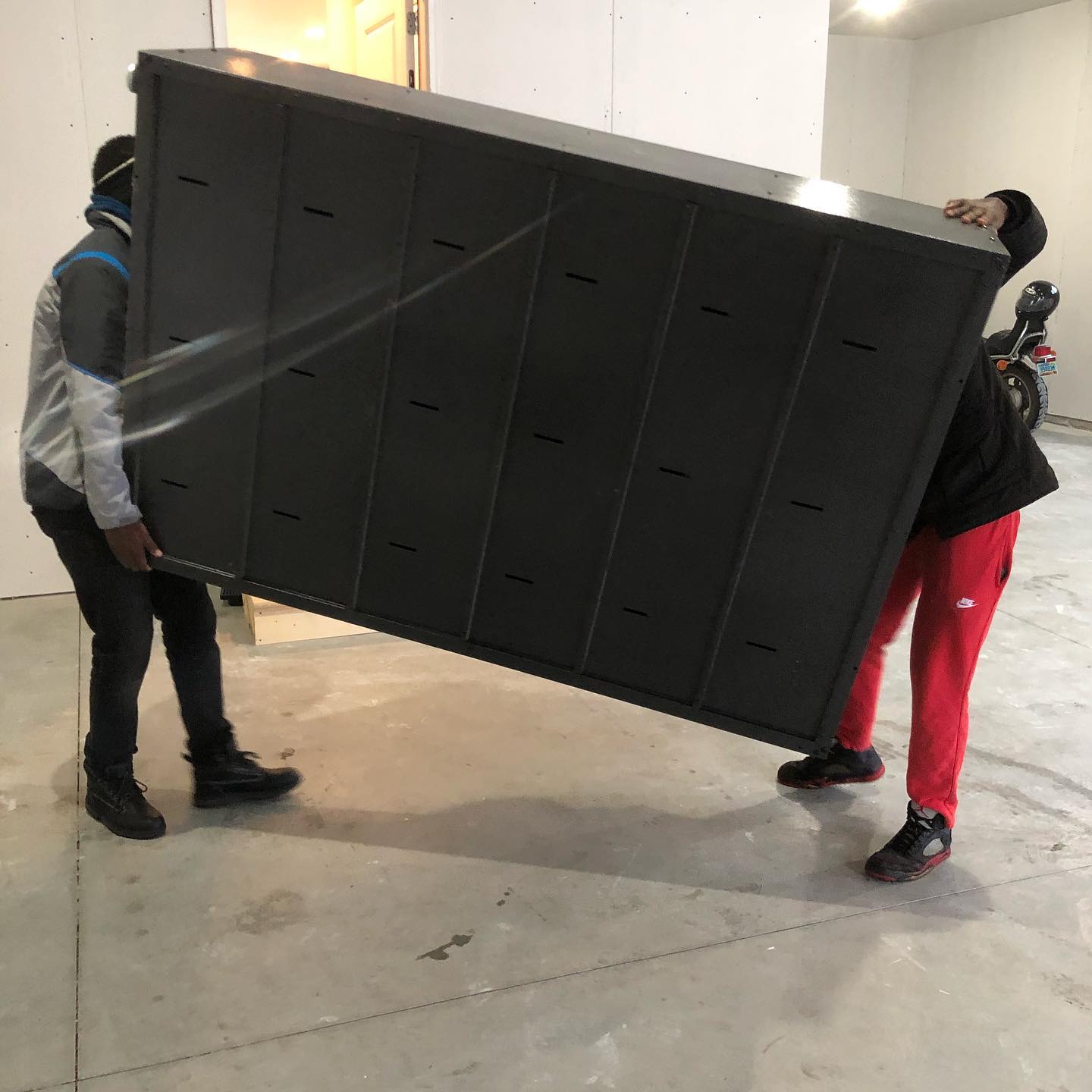 Two people carry a large, rectangular black locker unit through a room with a concrete floor.