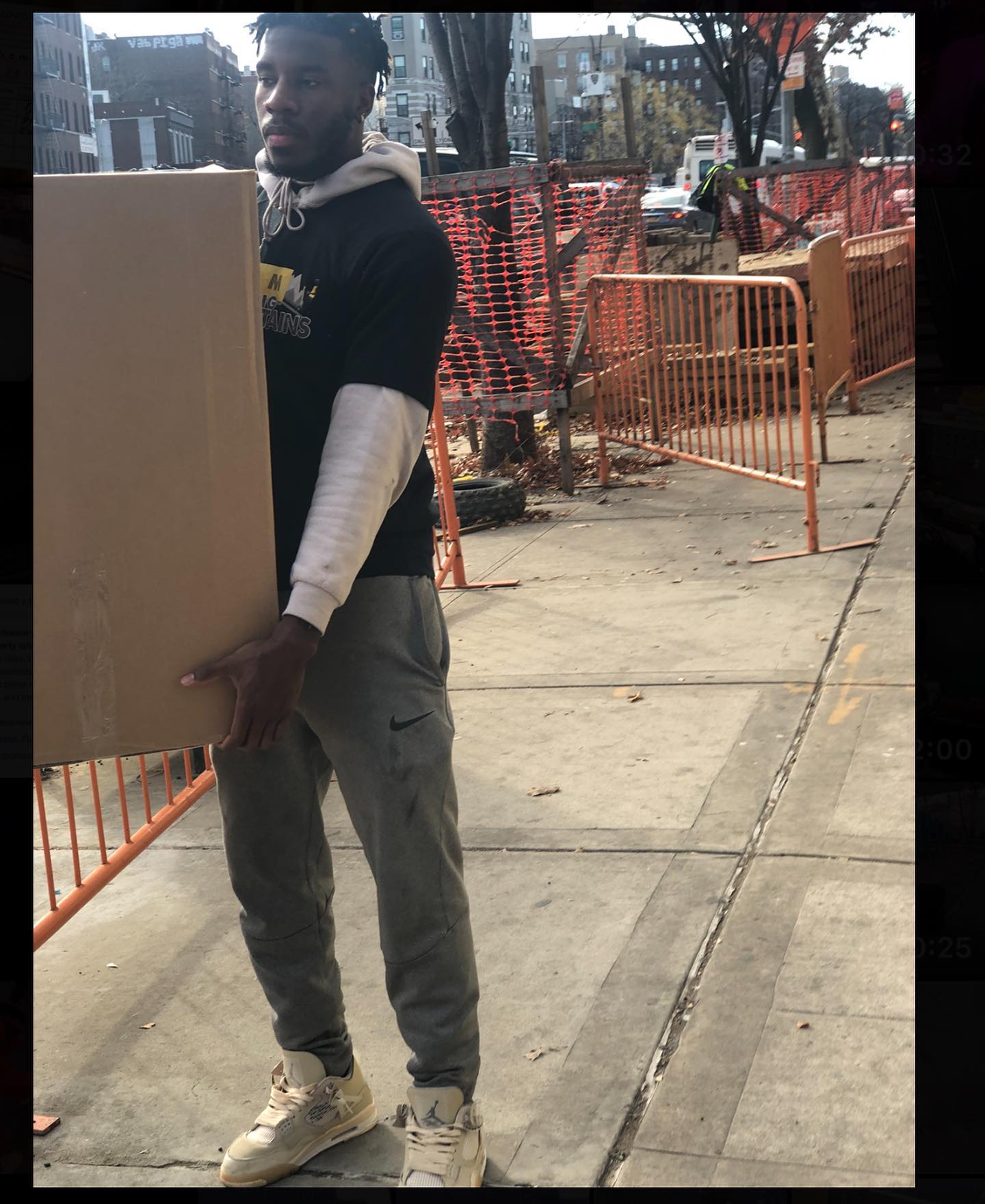 A person in a black shirt and gray sweatpants walks down a city sidewalk while carrying a large cardboard box.
