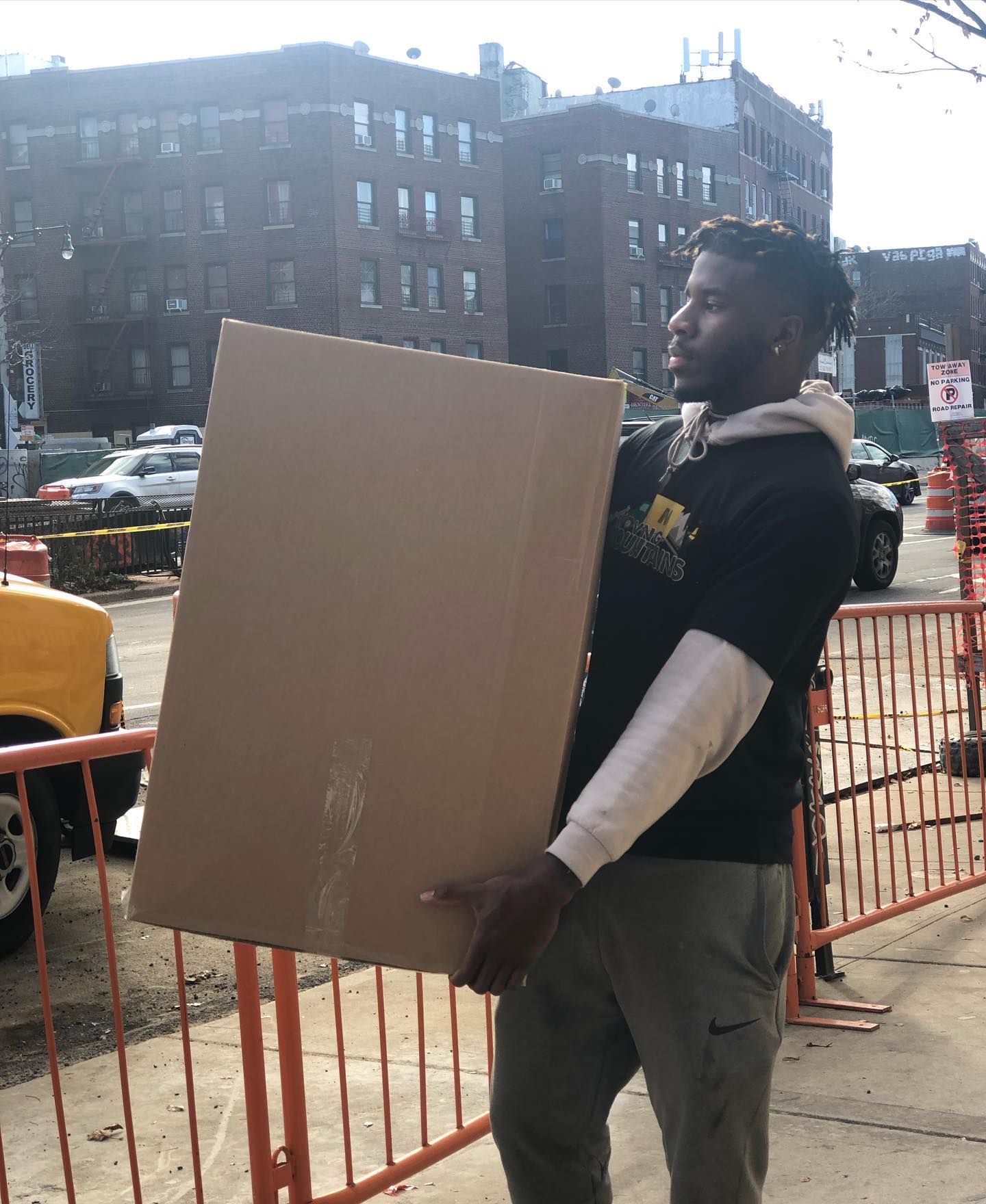 A person wearing a black hoodie and grey pants stands on a sidewalk, carrying a large cardboard box near an orange fence.
