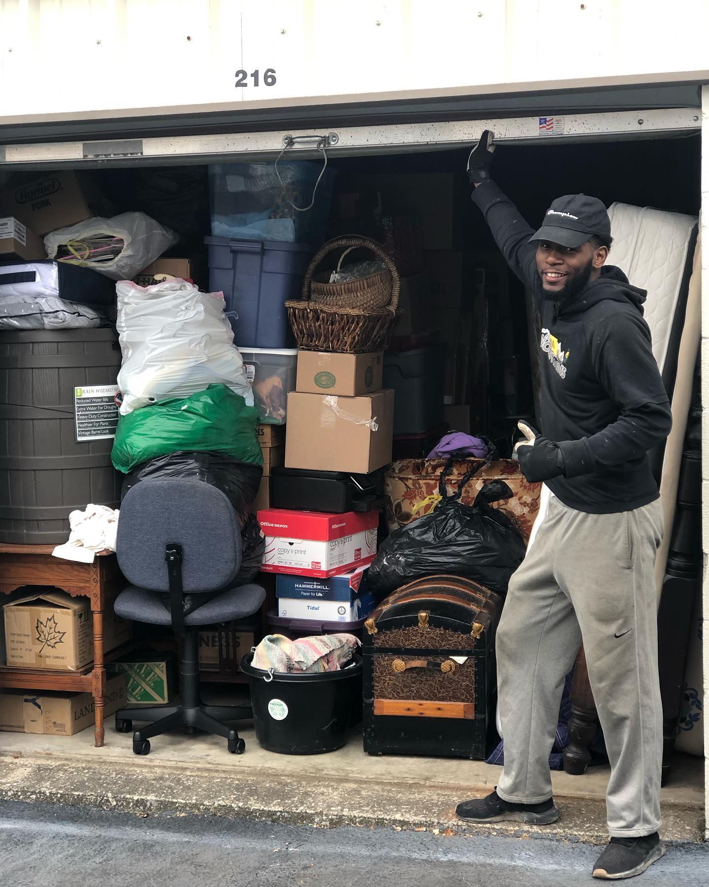 A person in a black hoodie and cap stands outside storage unit 216, smiling and leaning against the packed interior.