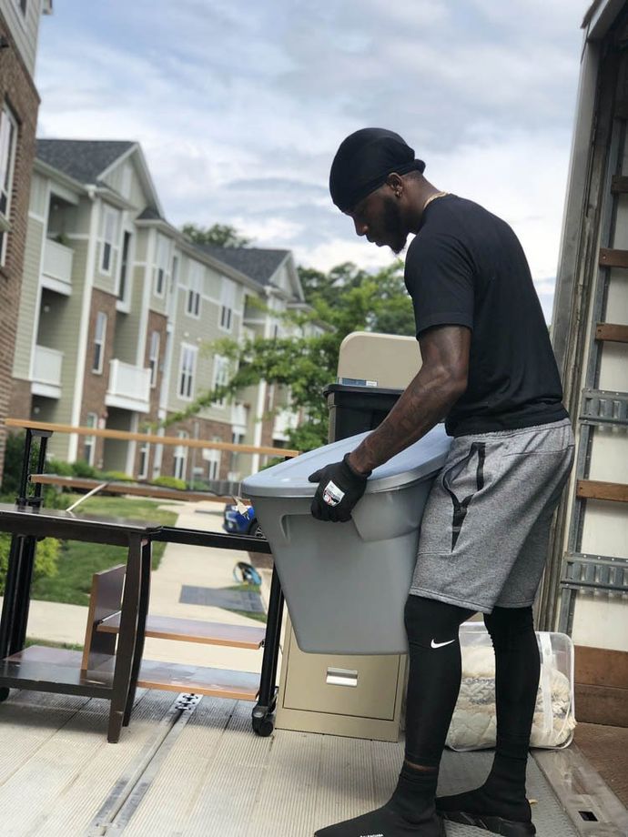 A person wearing a black shirt, shorts, and leggings carries a large gray plastic storage bin out of a moving truck.
