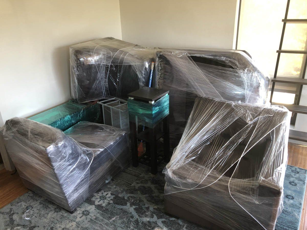 Furniture wrapped in clear plastic film sits on a patterned rug in a room with hardwood floors.