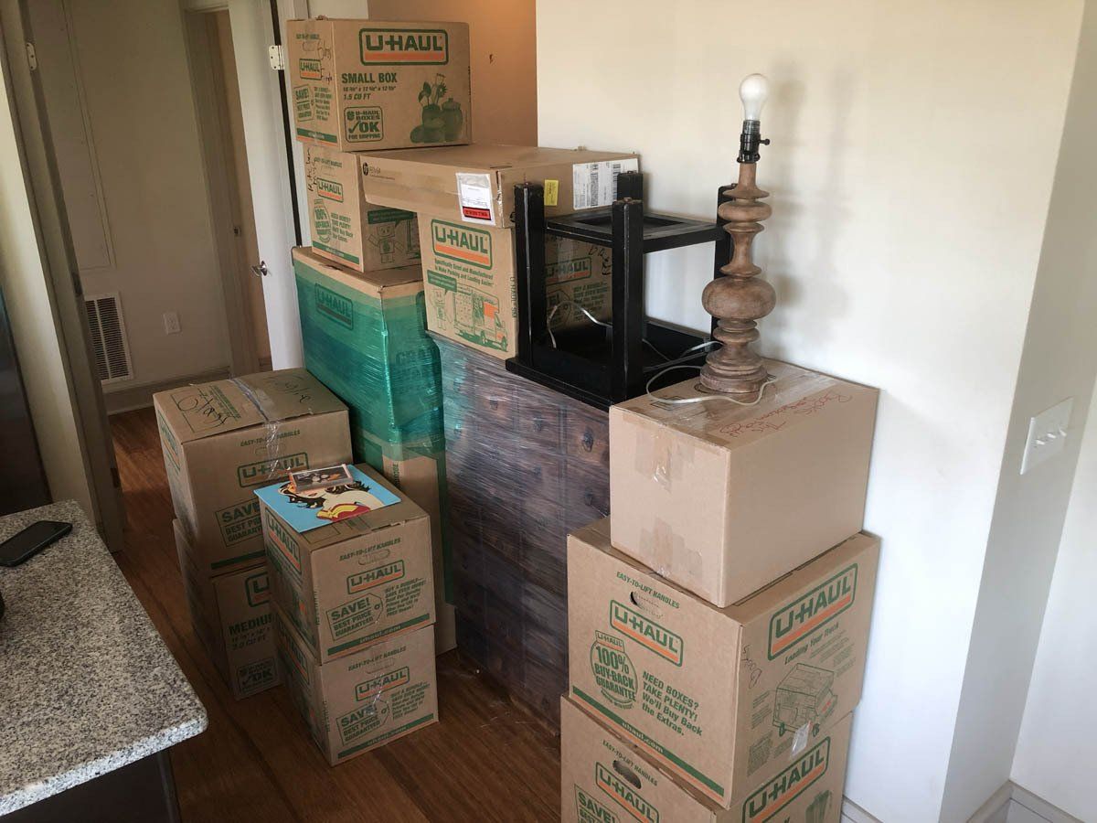 Stacks of cardboard moving boxes and a table lamp on a wooden floor in a room.