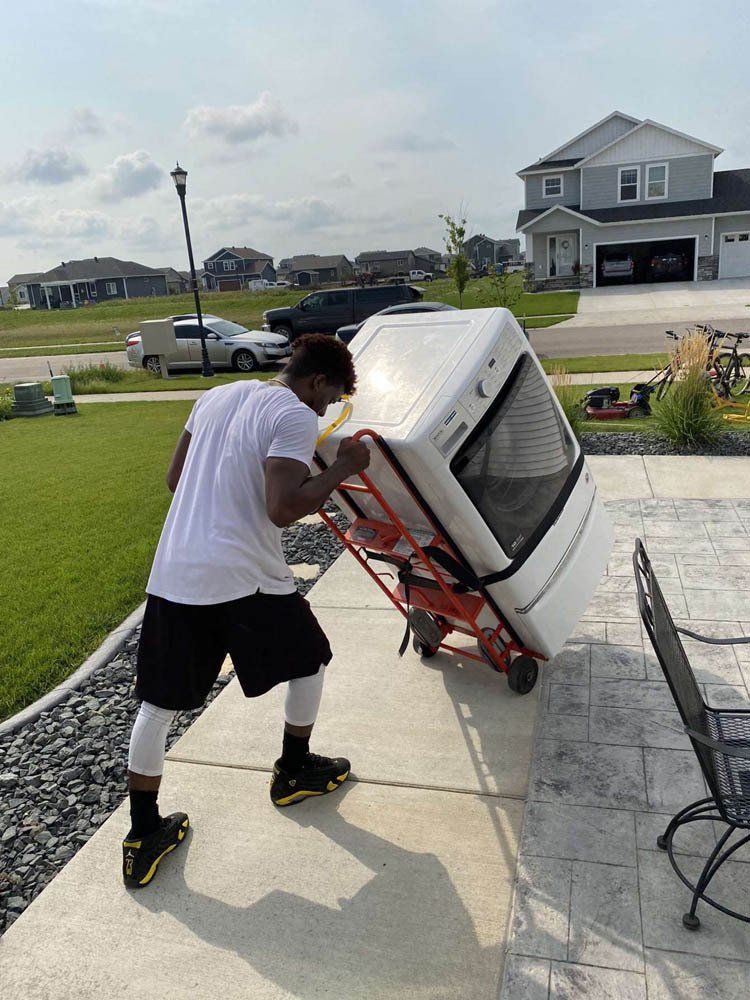 A person in athletic gear uses a red hand truck to move a white oven across a residential driveway.