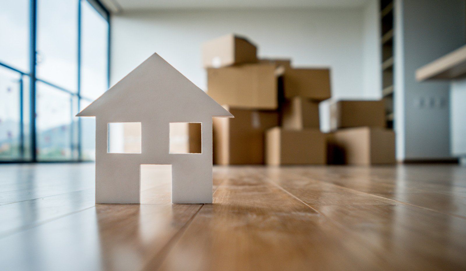 A small white paper cutout house stands on a hardwood floor in front of several stacked cardboard moving boxes.