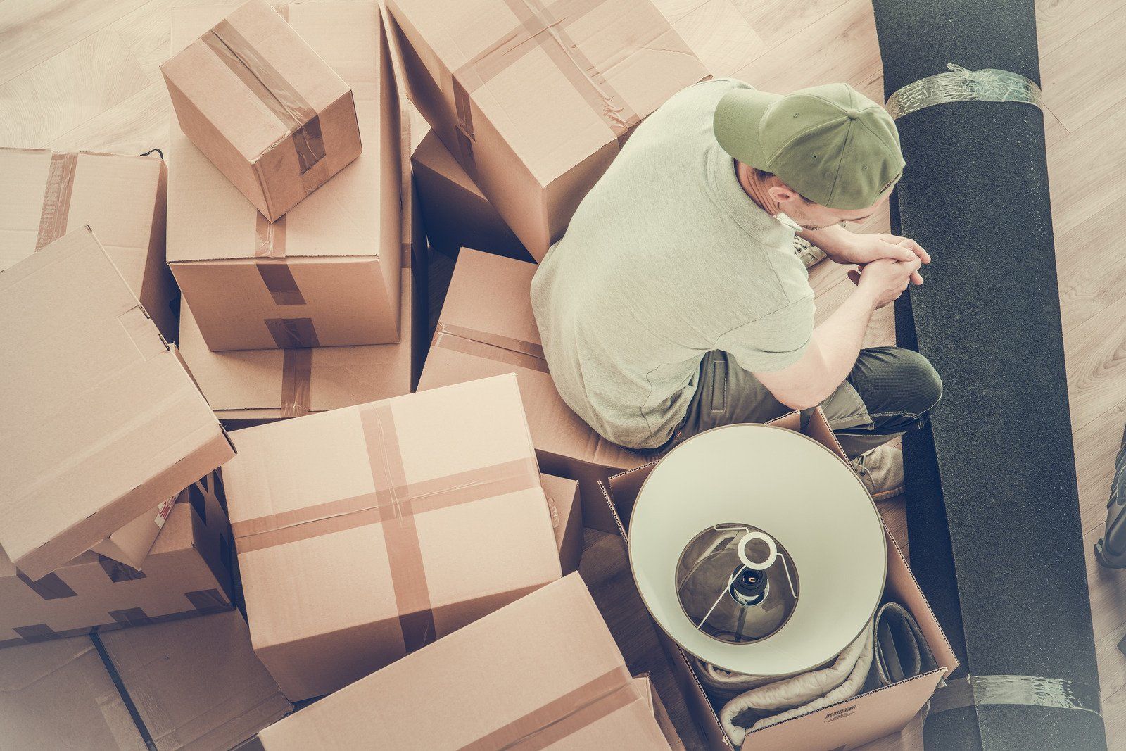 A person in a green cap sits on the floor amidst cardboard moving boxes, a lamp shade, and a rolled rug.