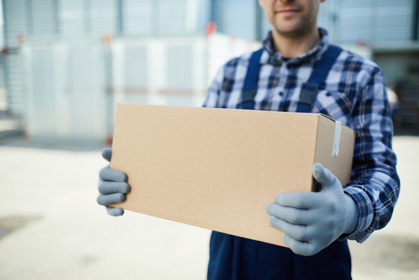A person wearing a plaid shirt and gray gloves holds a cardboard shipping box in an industrial warehouse setting.