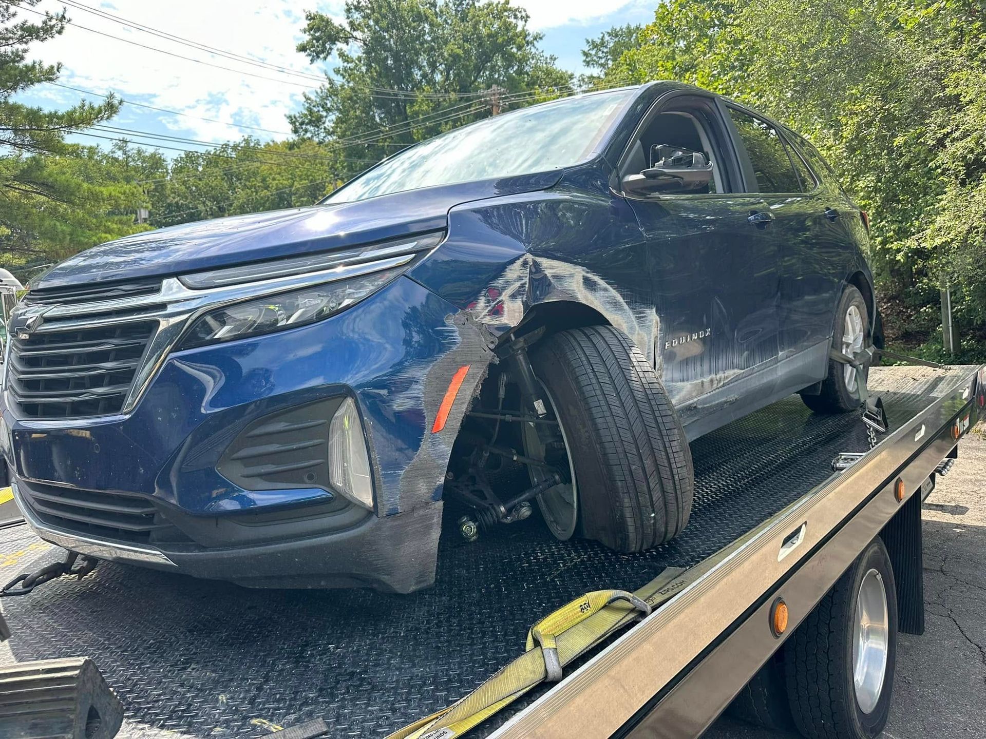 Blue SUV with front-end damage being towed by a truck on a sunny day.