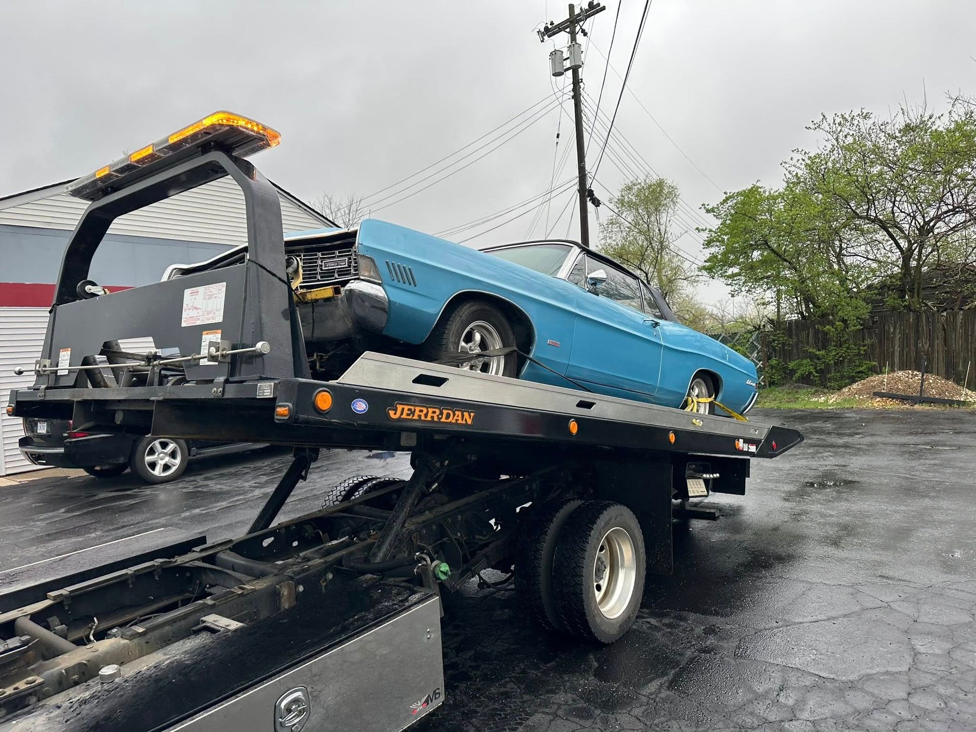 A light blue convertible being towed on a flatbed tow truck. Outdoors, overcast sky.