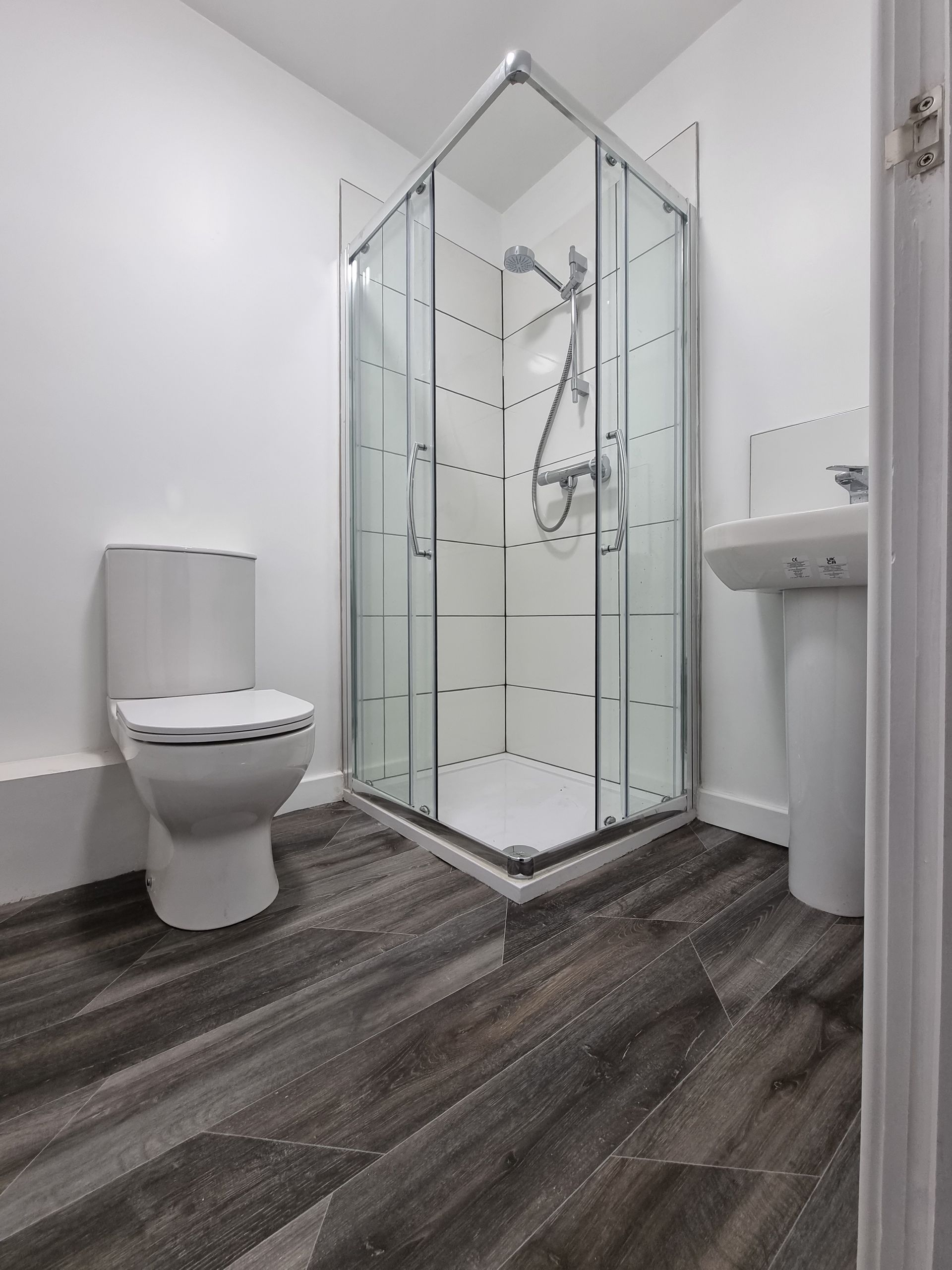 Small bathroom with toilet, corner shower, and pedestal sink. Dark wood-look flooring and white walls.