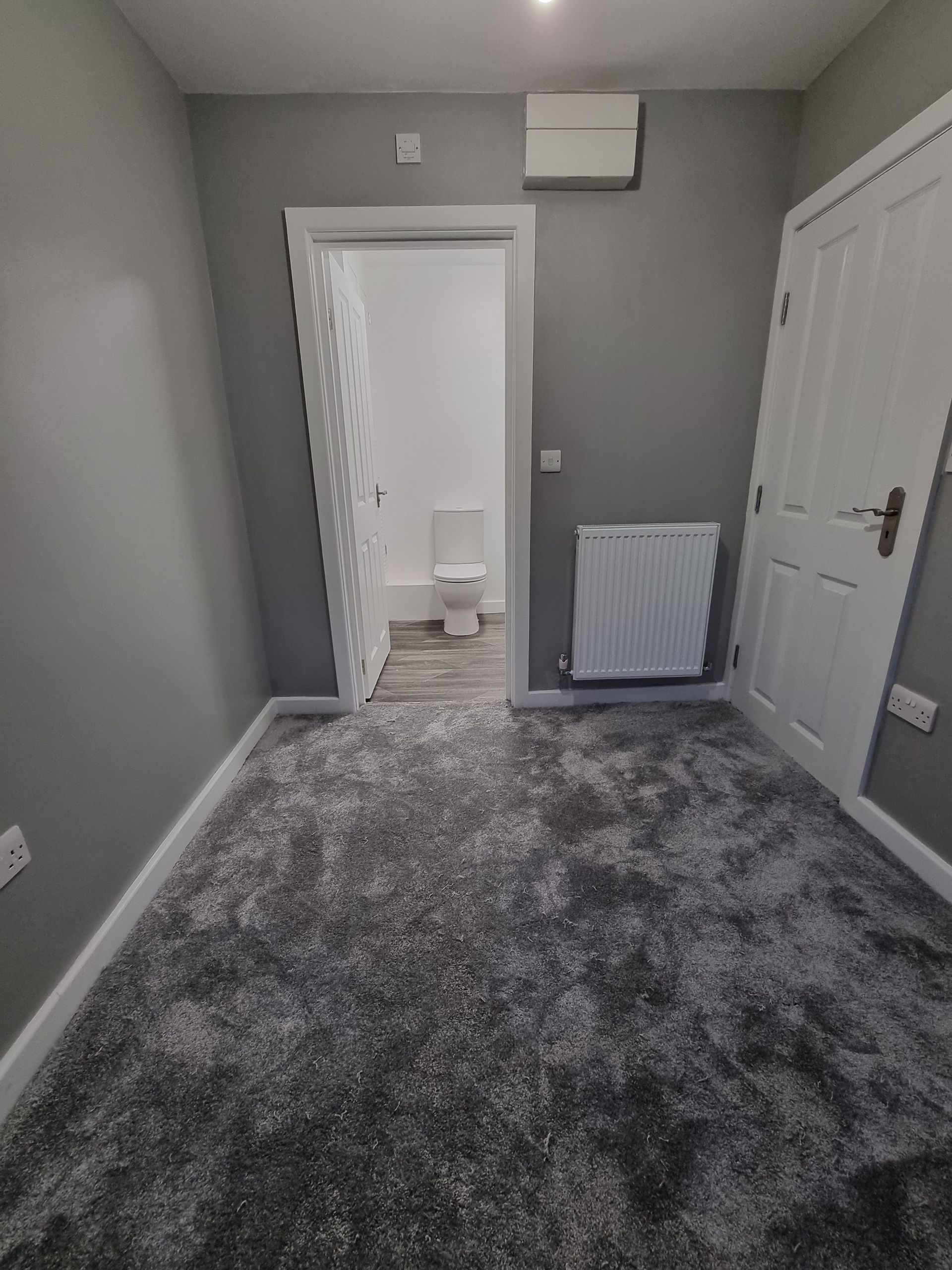 Gray-walled room with gray carpet and a white door leading to a bathroom with a toilet.
