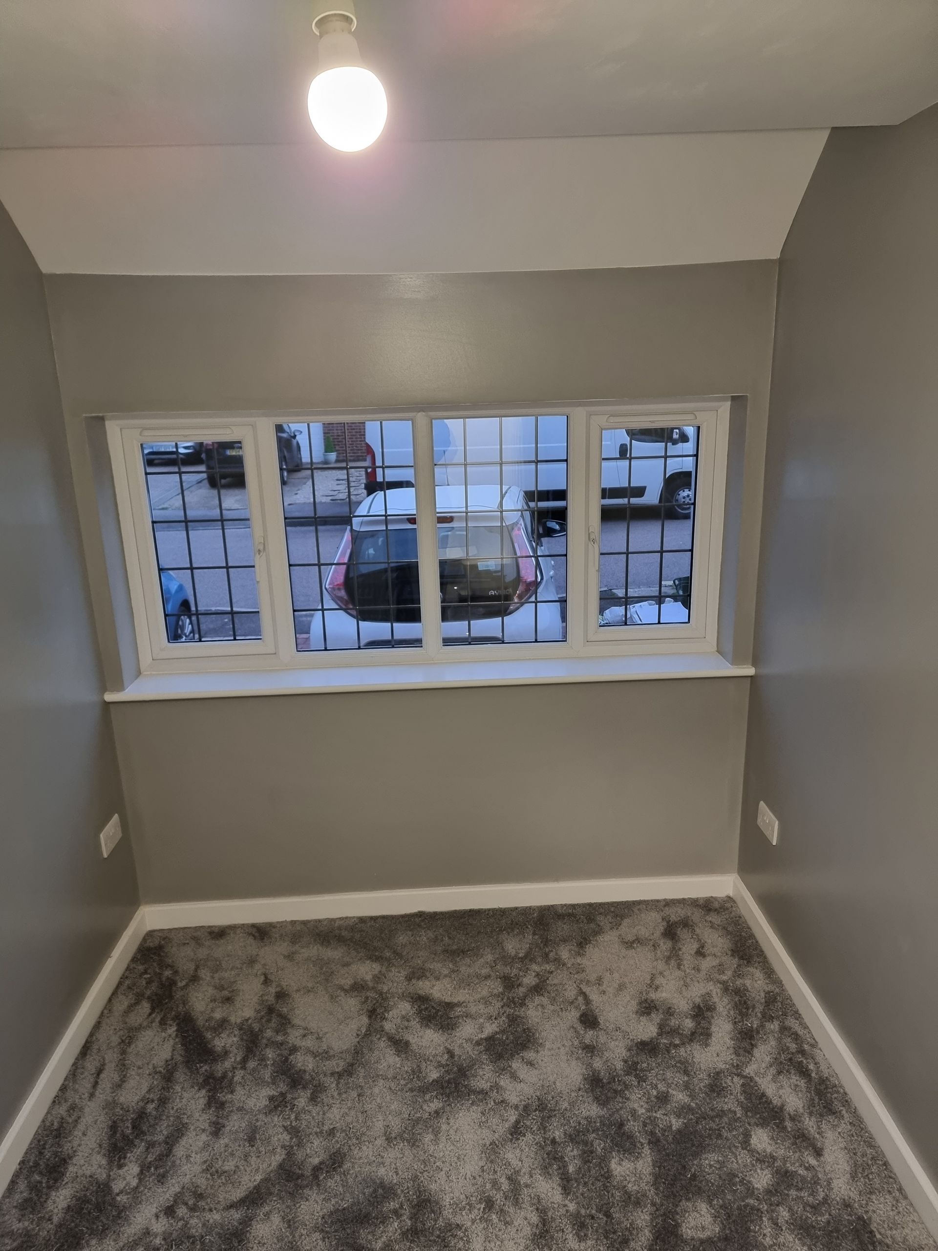Gray room with window overlooking a street. White trim, gray walls, and plush carpet. Light fixture on ceiling.