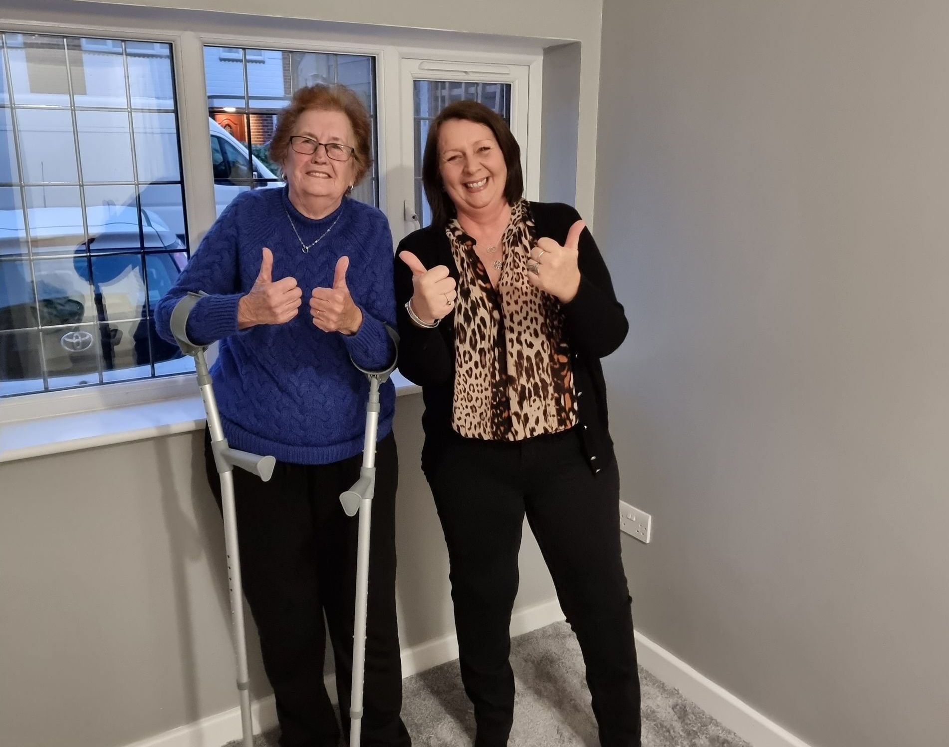 Two women giving thumbs-up in a newly renovated room. One uses crutches. Gray walls, window, and flooring.