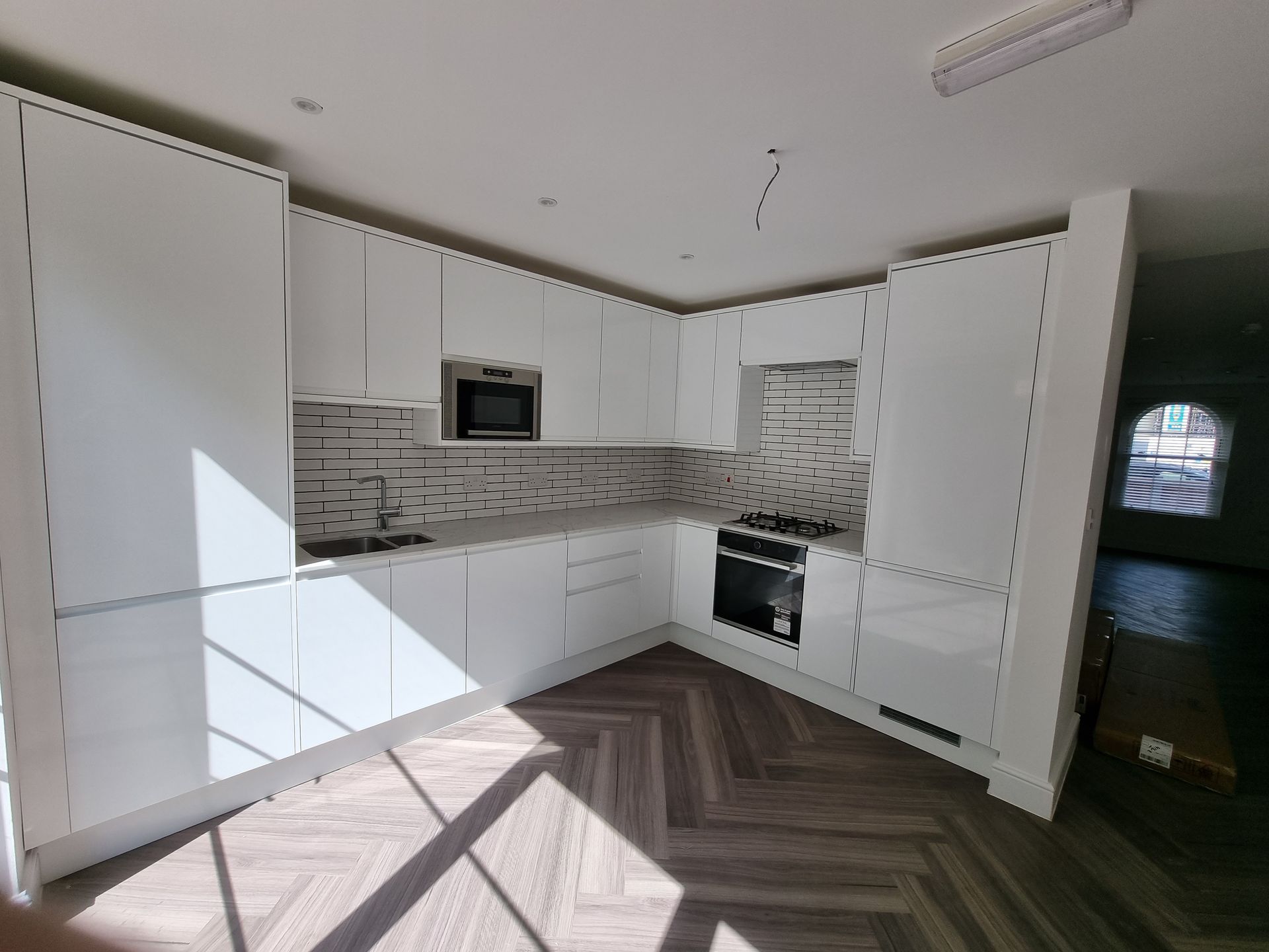 Modern white kitchen with herringbone floor. Cabinets, microwave, and oven are visible. Natural light.