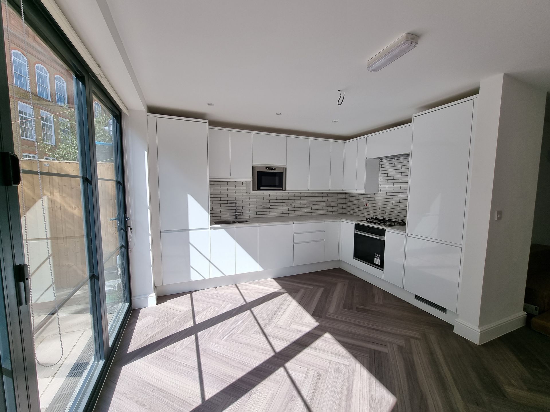 Bright, white kitchen with glossy cabinets, built-in appliances, and herringbone wood floor. Large glass doors to the left.