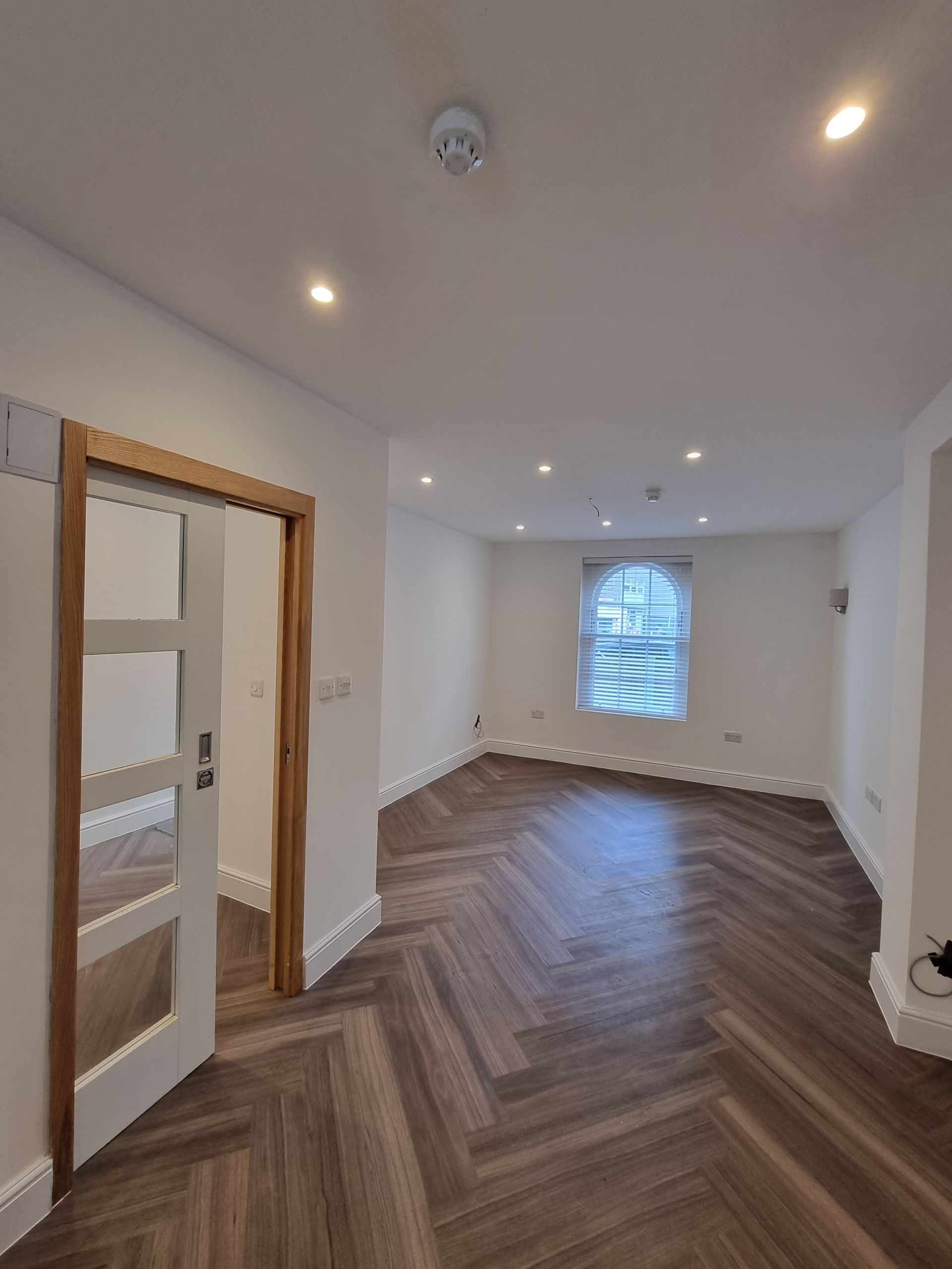 Empty room with wooden herringbone floors, white walls, a doorway, and a window.