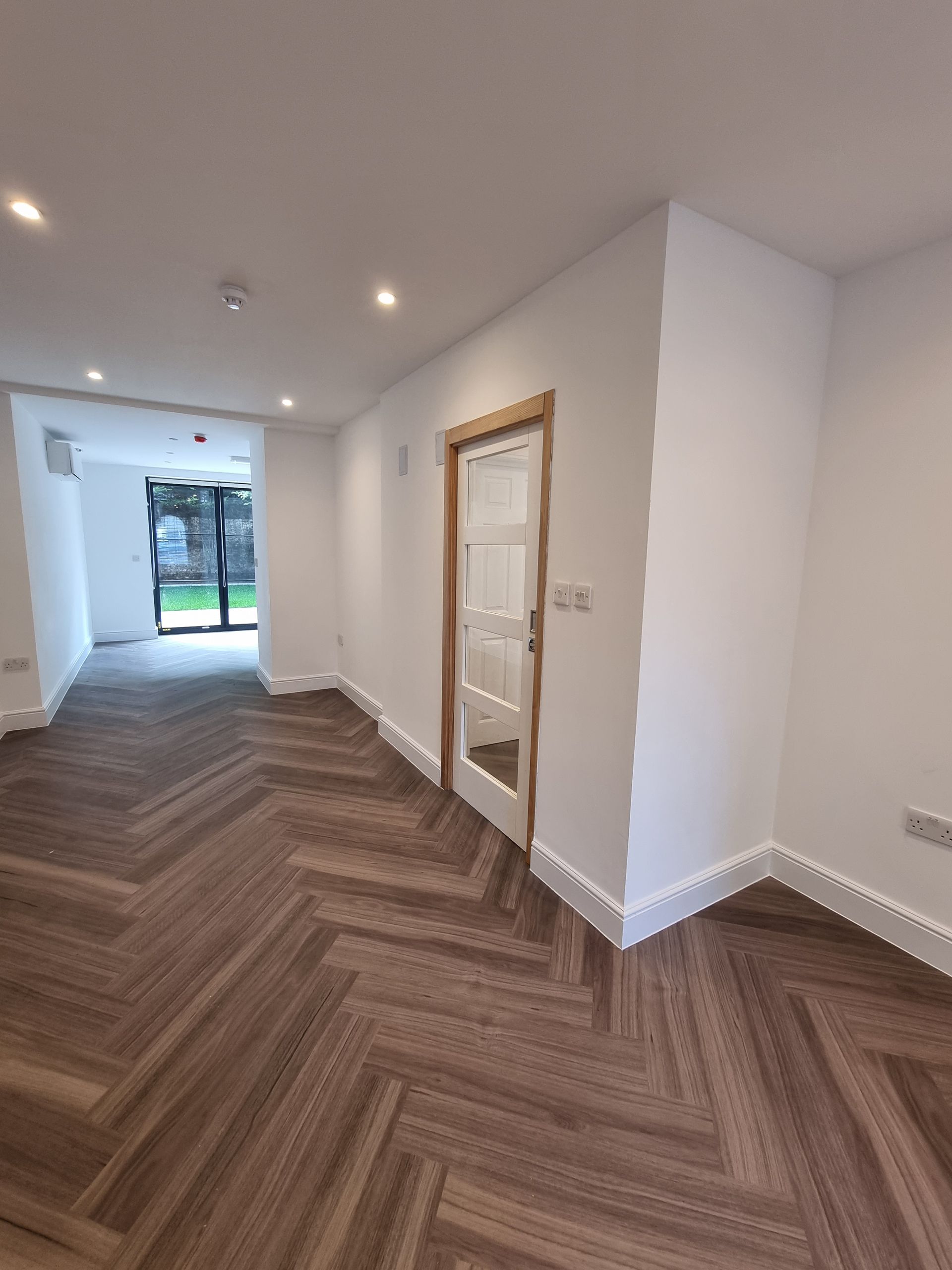 Interior of a room with herringbone wood flooring, white walls, and a glass-paneled door.