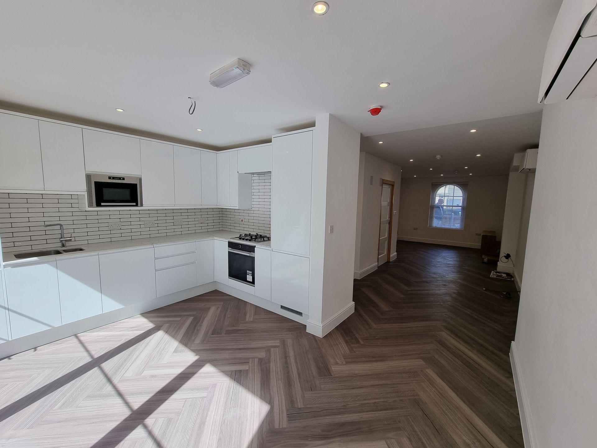 Bright, modern kitchen with white cabinets, herringbone wood floor. View into adjoining room with window.