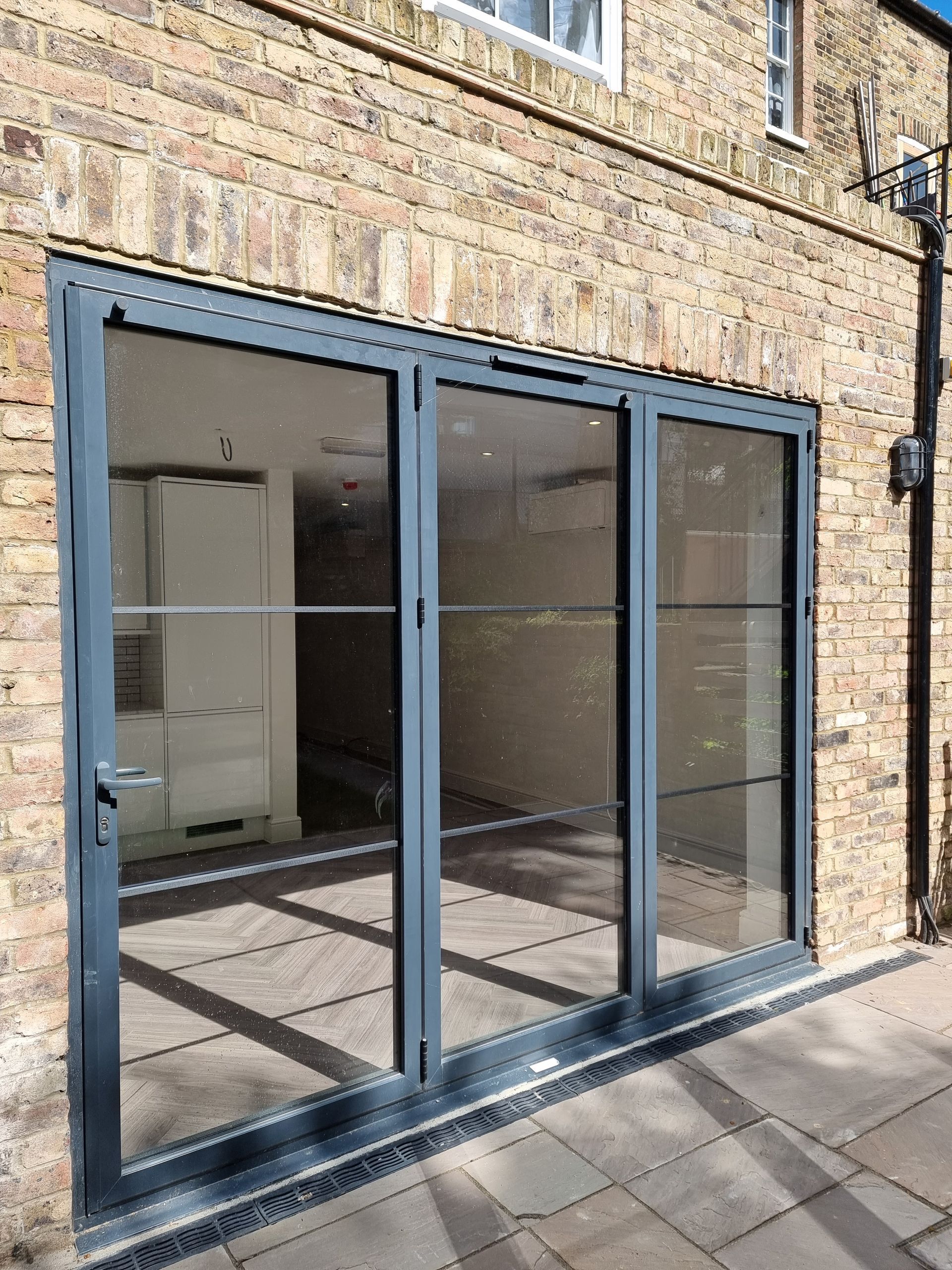 Three-panel glass door with dark gray frame in a brick wall. Sunlight reflects on the glass.