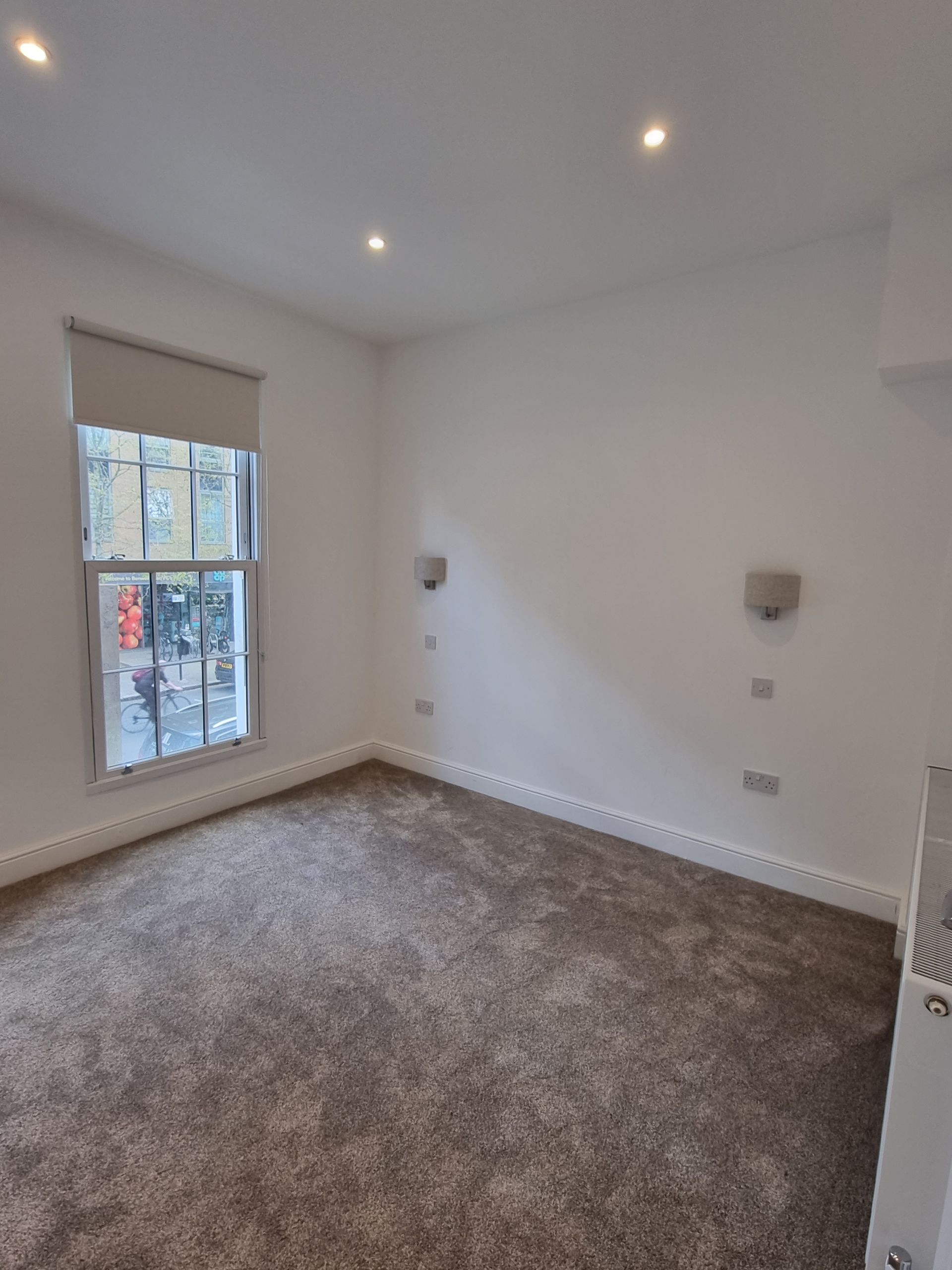Empty room with window, carpet, and white walls. Two sconces and electrical outlets visible.