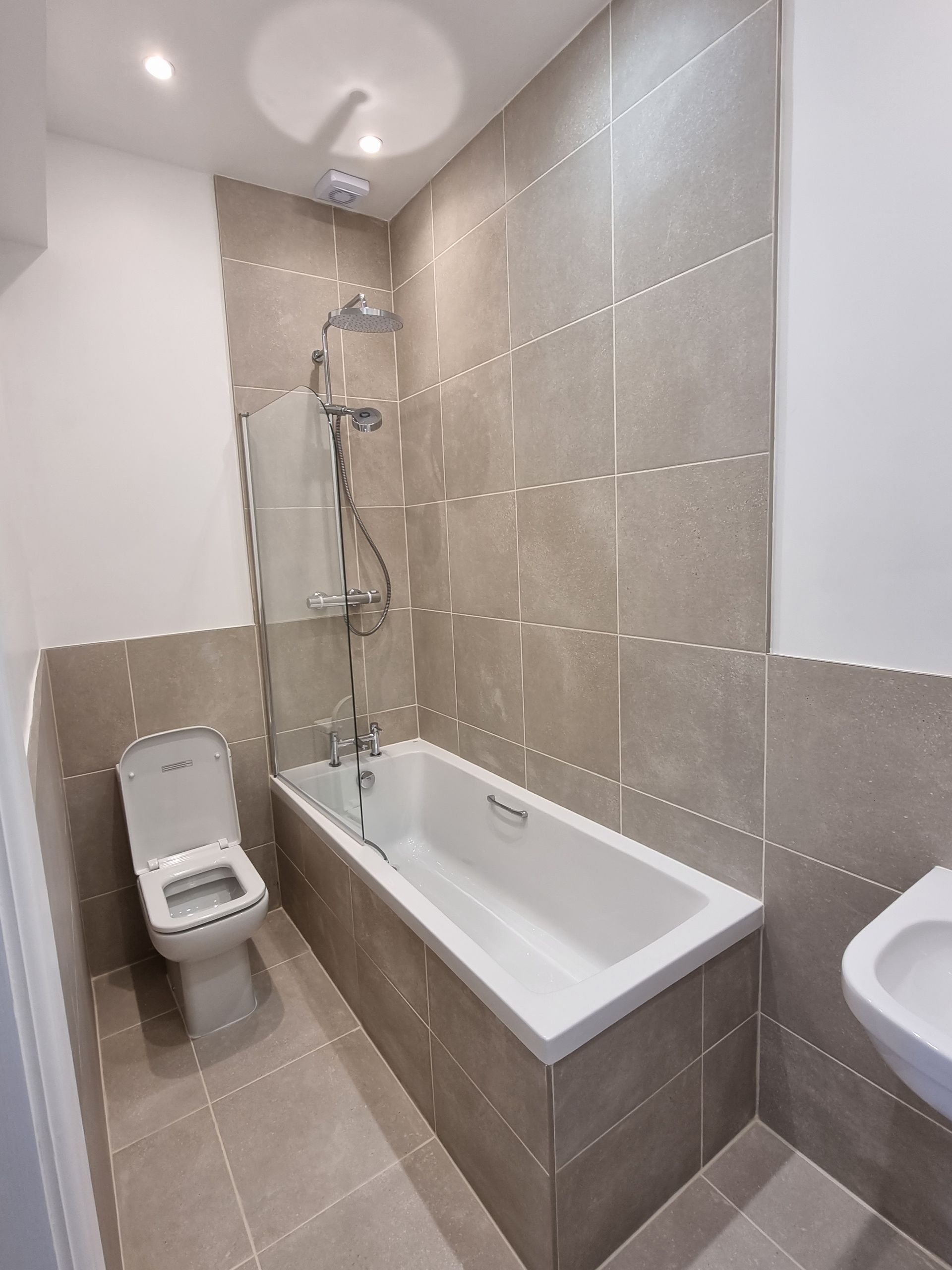 Small modern bathroom with a bathtub, toilet, and sink. Beige tile walls and flooring.