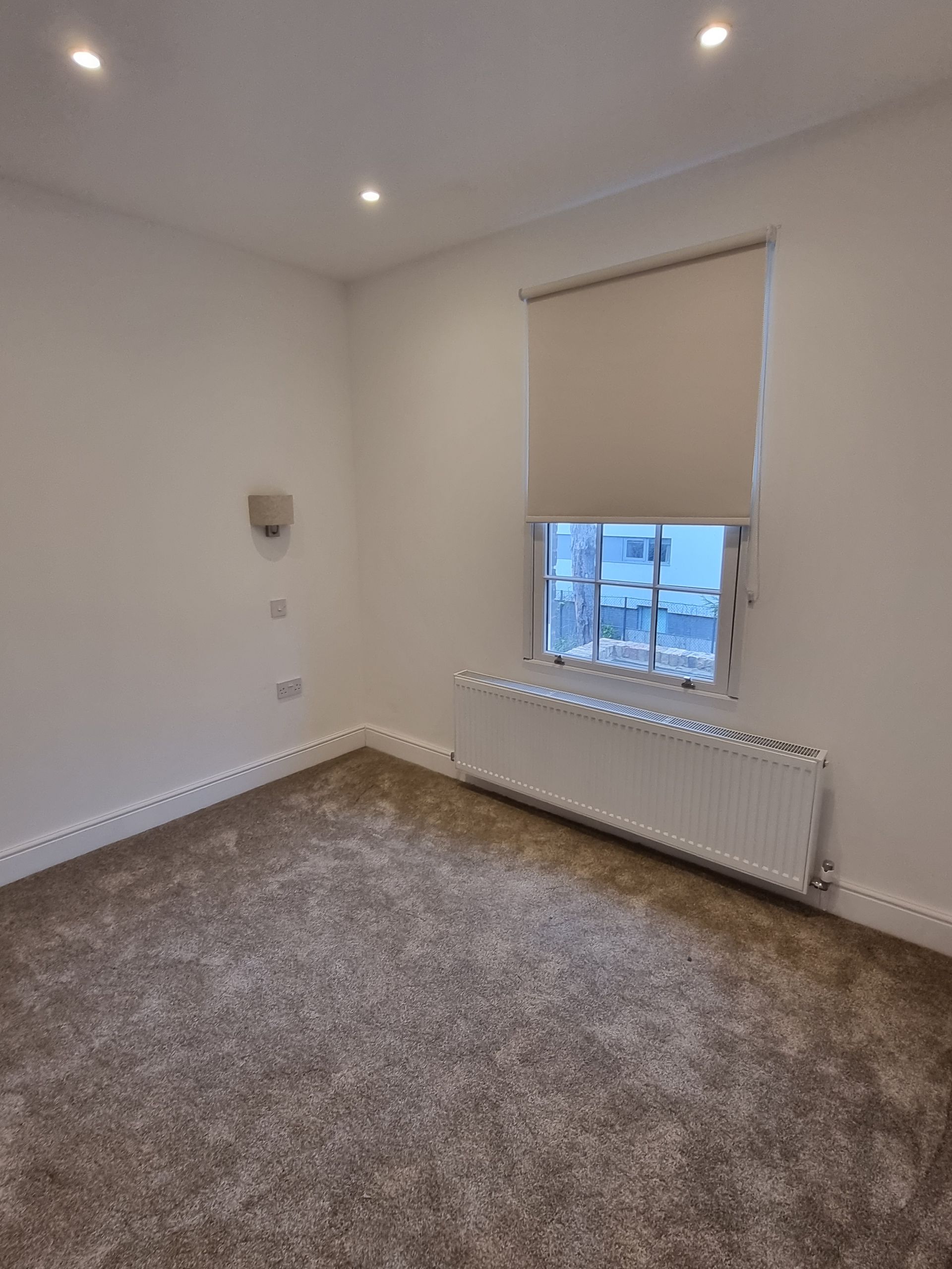 Empty room with beige walls, brown carpet, window with closed blind, and radiator.