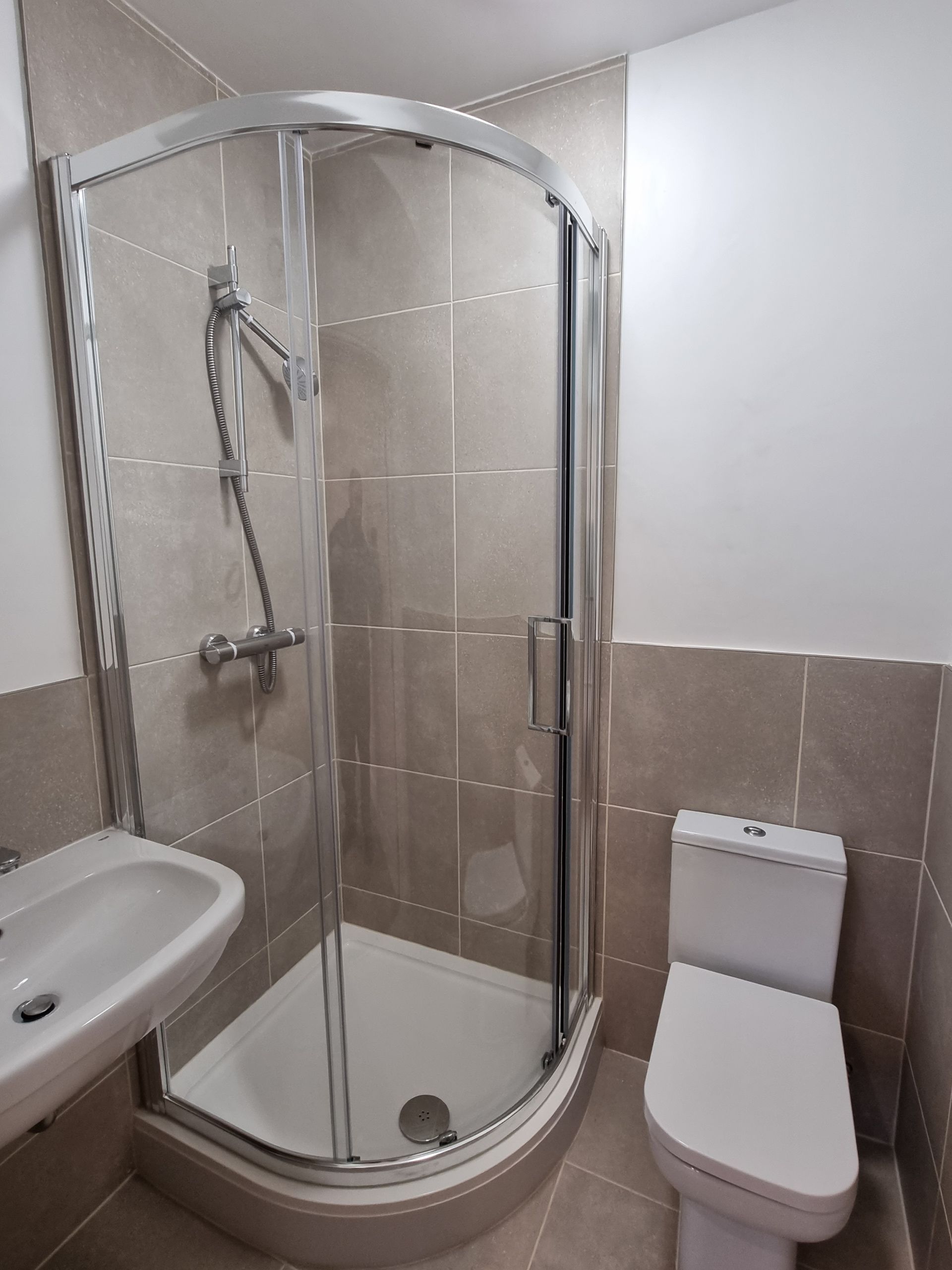 A small bathroom with a curved glass shower, sink, and toilet; beige tile walls.