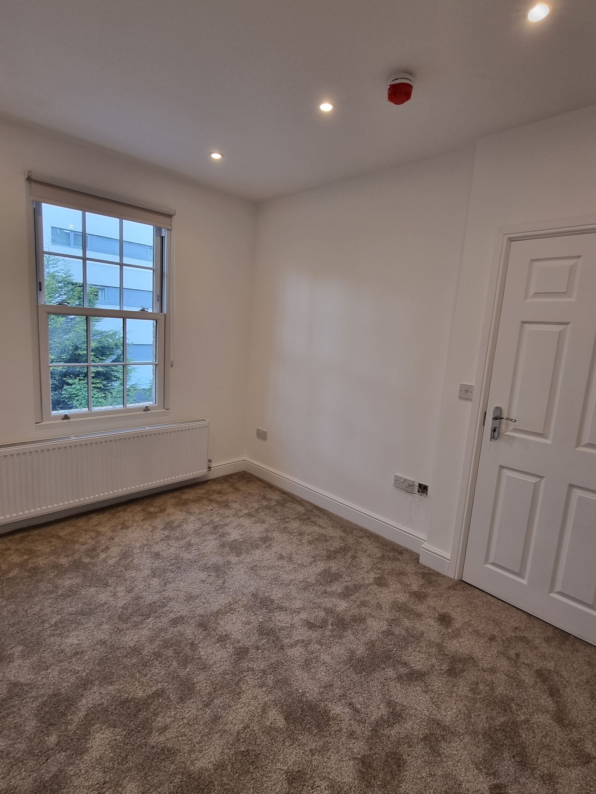 Empty room with a window, white walls, carpet, and a closed door.