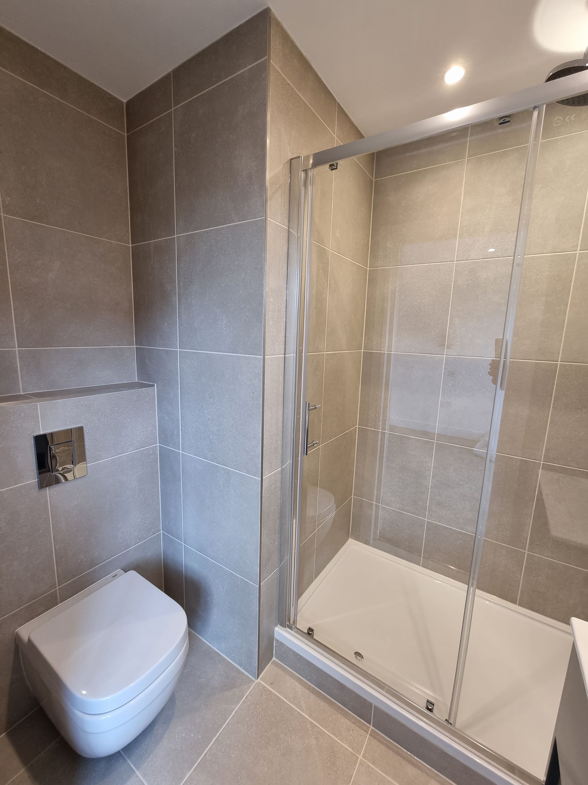 Bathroom with gray tiled walls, a toilet, and a glass-enclosed shower.