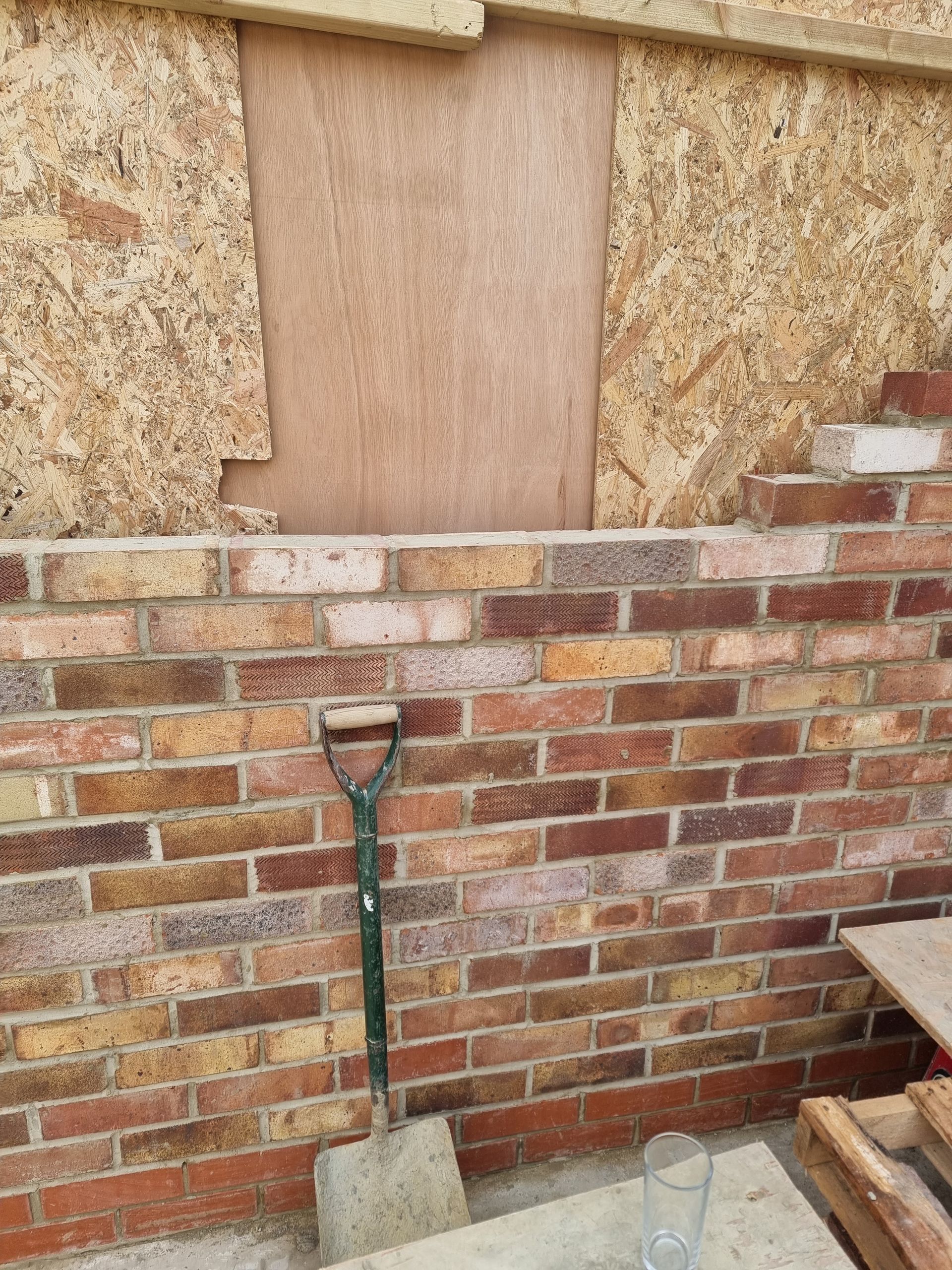 Brick wall under plywood, shovel leaning against it.